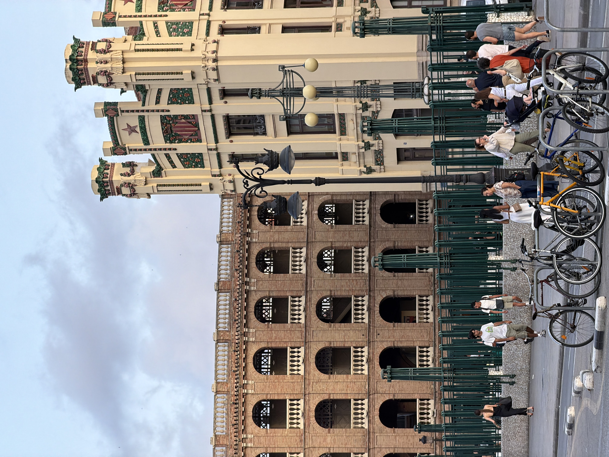 The image depicts an urban street scene in front of two distinct architectural structures. On the left, a large, multi-story building constructed of reddish-brown brick features a repeating pattern of arched windows and balconies with white balustrades. Green, vertical metal posts create a fencing or barrier in front of this building. To the right, a cream-colored building with ornate architectural details, including decorative motifs, star symbols, and shields, stands prominently. This building has multiple windows and appears to be several stories high. In the foreground, several bicycles are parked. People are walking on the sidewalk, some interacting with each other, while others are engaged in individual activities. The sky above is partly cloudy. The overall impression is a busy city environment with notable historical or distinctive architecture.