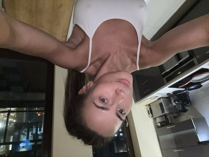 The image displays a woman taking an inverted selfie indoors, presenting a unique, upside-down perspective.
The primary subject is a woman with long, dark hair, which falls downwards (towards the top of the frame in the current orientation). Her face, positioned centrally, looks directly into the camera with a neutral to slightly smiling expression. Her eyes are open and a light color, possibly green or hazel. Her facial features, including her nose, lips, and eyebrows, are clearly visible. She has a clear complexion. She is wearing a simple white tank top or sports bra with thin straps, which appears somewhat fitted and slightly sheer in places. Her shoulders, upper chest, and arms are visible, showing muscle definition.
The background behind the woman is split into two main sections:
On the left side (when viewing the image upright), a large window or glass door is visible, looking out into a dark night. Various lights from outside, likely from buildings or streetlights, are visible through the glass, indicating an urban or suburban setting after dark. The window has a dark frame.
On the right side (when viewing the image upright), a modern kitchen area is visible. There is a light-colored countertop, possibly white or light grey, extending along the wall. On the countertop, a black stovetop with a red-bottomed frying pan is present. Below the countertop, built-in appliances, including a dark oven or microwave, are visible. Further along the counter, another kitchen appliance, possibly a blender or food processor, is also seen. The cabinetry below the countertop appears to be dark grey or black, contrasting with the lighter countertop and floor. A light-colored tiled floor is partially visible towards the upper right of the frame (when upright).
The overall impression is a casual, nighttime indoor scene, captured with an artistic or playful inverted selfie. The lighting suggests artificial indoor lighting, effectively illuminating the subject and the immediate surroundings.