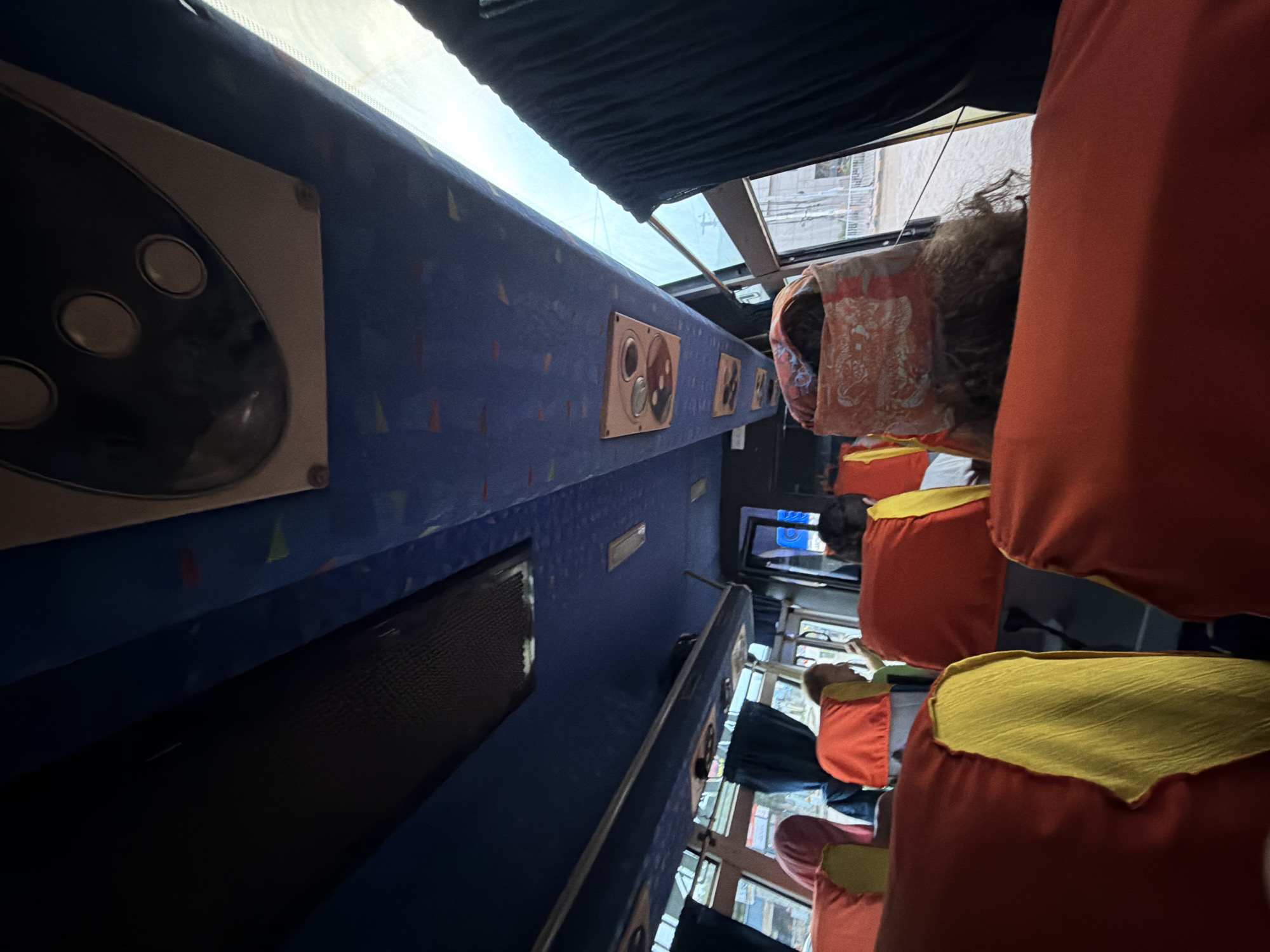 The image captures the interior of a bus, likely in Hyderabad, India, as indicated by the presence of a public bus's seating arrangement and visible exterior elements. The perspective is from a passenger's seat, looking forward and upwards. The bus interior is characterized by blue upholstery on the ceiling and overhead compartments. Red and yellow seat covers are visible in the foreground and middle ground, suggesting a public transport vehicle. Several passengers are seated, with their heads and upper bodies visible. One passenger in the foreground wears a patterned head covering. Outside the bus windows, a street scene is partially visible, with buildings and what appears to be overcast or potentially wet conditions. The lighting suggests it is daytime, possibly with diffused light due to weather.