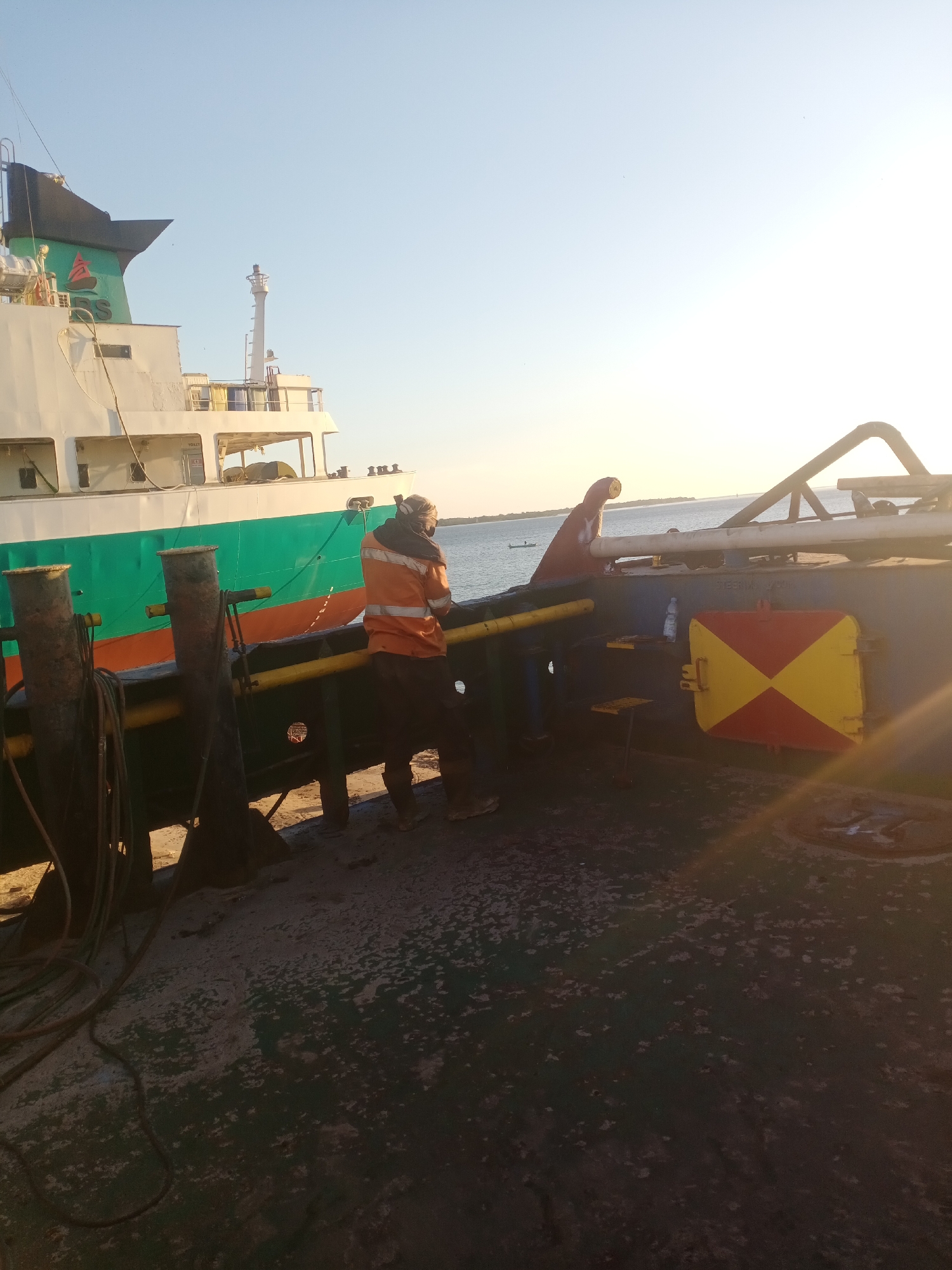 The image depicts a dockside scene at sunset. A person wearing an orange high-visibility jacket and dark pants is standing on a dark-colored platform, looking towards a large cargo ship. The ship has a white superstructure, a green and red hull, and a black funnel with a green emblem. The platform is equipped with bollards and ropes. In the background, the sea is visible under a clear sky with the sun setting. A red and yellow diamond-shaped marker is affixed to the side of a structure on the platform. The overall impression is one of an industrial setting during a calm evening.