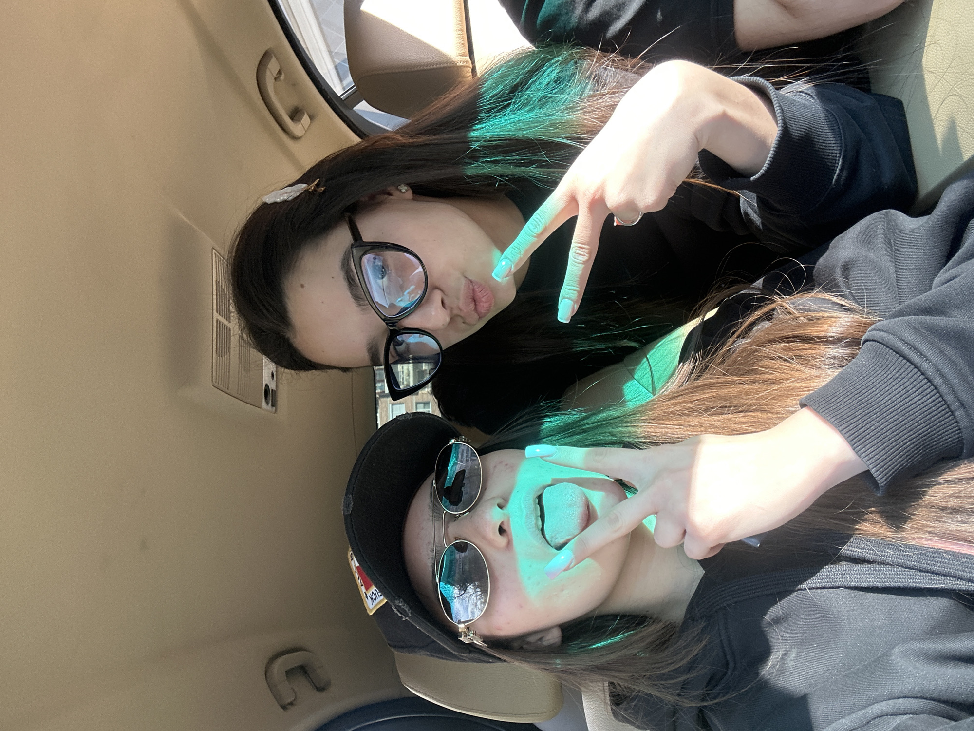 Two young women are posing for a selfie inside a car. The woman on the upper left wears glasses and is making a kissing face with a peace sign. The woman on the lower right has a baseball cap and sunglasses and is sticking out her tongue with a peace sign. They are both wearing black hoodies. The interior of the car is beige. No information in the image allows for location in Plovdiv, Bulgaria.