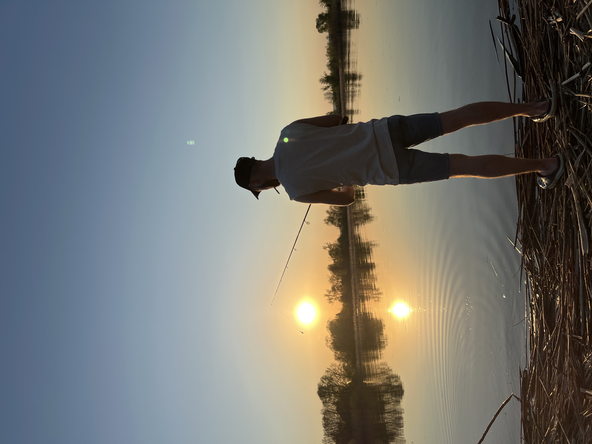 A man in a white t-shirt, grey shorts, a black baseball cap, and grey slip-on shoes is fishing at sunset. The man is holding a fishing rod and appears to be casting. The location is near a body of water, possibly a lake or river, near the city of Borovoye Matyushino, Russia. Reeds are present on the shoreline. The sunset is reflected in the water.