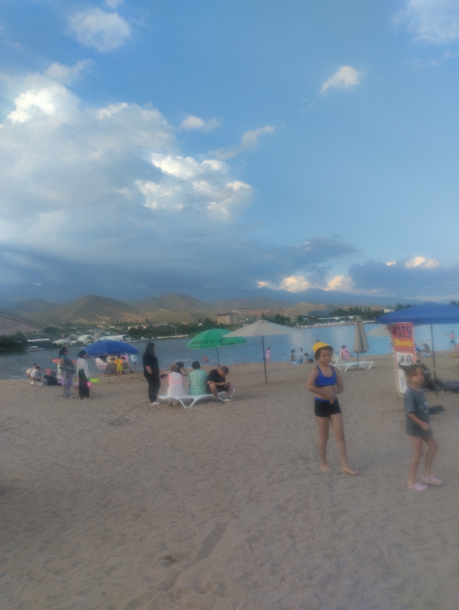 This image captures a scene at a beach in Cholpon-Ata, Kyrgyzstan, on a cloudy day with a bright sky. The foreground is dominated by a sandy beach where several people are gathered. In the mid-ground, a body of water, likely Lake Issyk-Kul, is visible, with a town and mountains forming the backdrop.
Several individuals are present on the beach. Two young girls are prominently featured in the right side of the frame. One, wearing a yellow hat and a blue swimsuit, stands facing forward. The other, to her right and slightly behind, is in a gray t-shirt and dark shorts. Further back, a group of people are seated on beach chairs under umbrellas. To the left, several adults and children are seen walking and relaxing on the sand.
Various beach amenities are scattered across the scene. Umbrellas, some blue and some green, provide shade. Beach chairs are visible, some occupied and some empty. In the background, across the water, a town with buildings and resort-style structures can be observed, nestled at the foot of rolling hills that transition into imposing mountains. The water itself appears calm, reflecting the cloudy sky. The overall atmosphere suggests a leisurely day at a popular tourist destination.