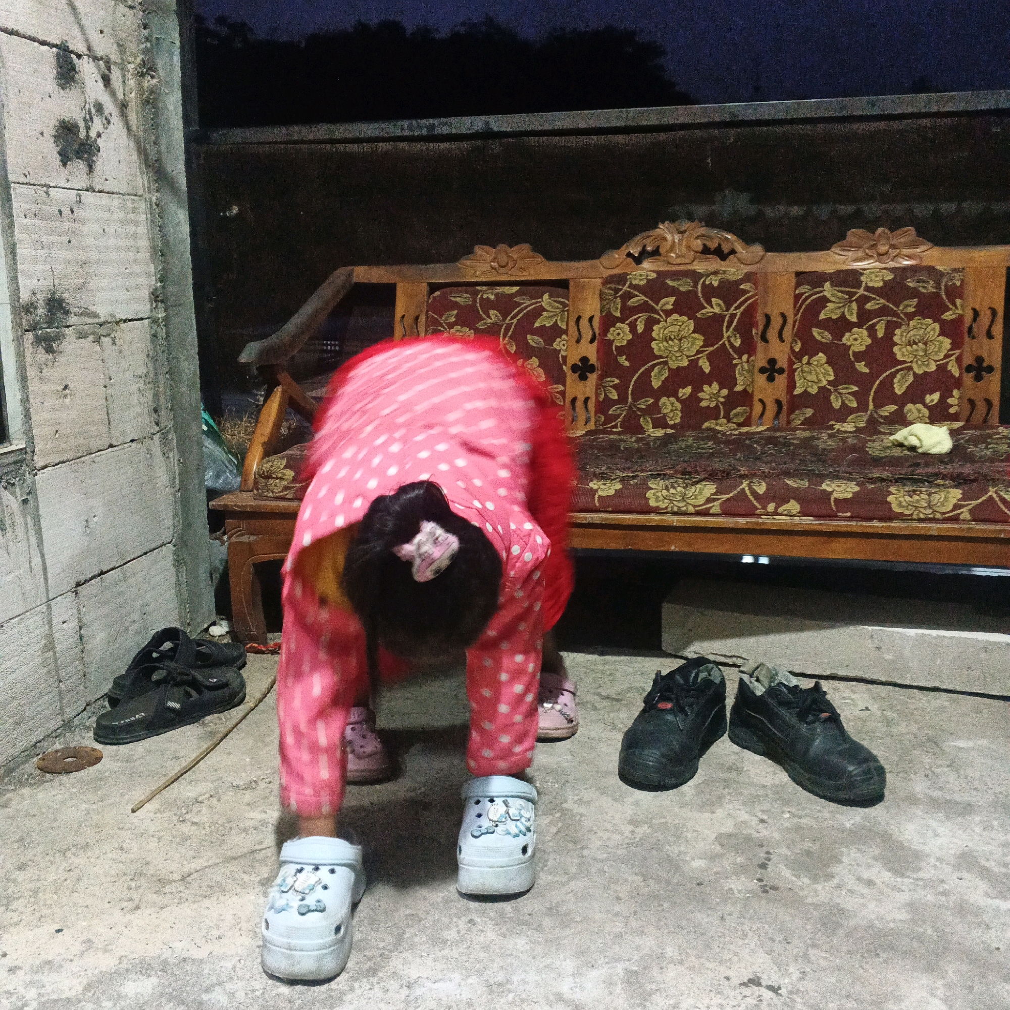 A child, wearing a pink polka dot shirt and pants, is in a bent-over position on a concrete floor. The child's face is not visible. A pair of light blue clogs with cartoon characters are on the child's feet. To the left of the child, a pair of black sandals are on the ground. To the right of the child, a pair of black work boots are on the ground. In the background, a wooden bench with a floral pattern upholstery is visible. The scene appears to be outdoors, possibly a backyard or patio, in Bocek, Indonesia. The lighting suggests it is dusk or evening.