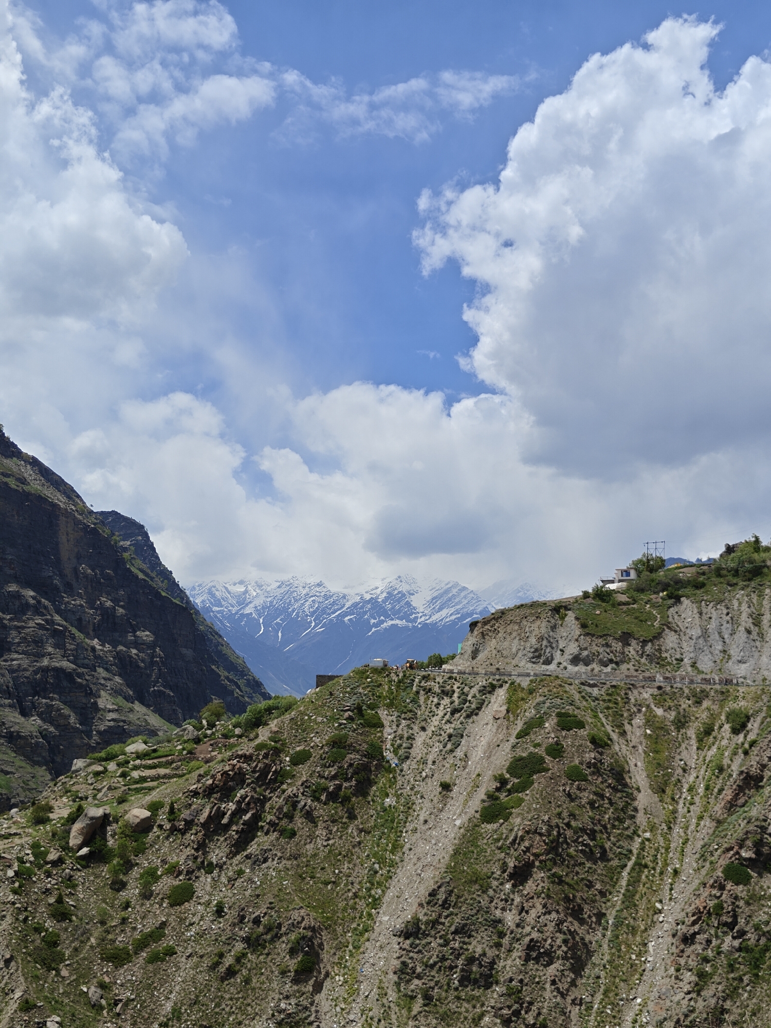 The image presents a mountainous landscape in Shuiling, India, under a partly cloudy sky. In the background, snow-capped mountains are visible. A steep, rocky hillside with sparse vegetation dominates the foreground. A narrow road with a few vehicles winds along the side of the hill. On a ridge above the road, a small building with utility infrastructure is perched. To the left, a dark, sheer cliff face rises, contrasting with the lighter slopes on the right. The sky is blue with scattered white clouds.