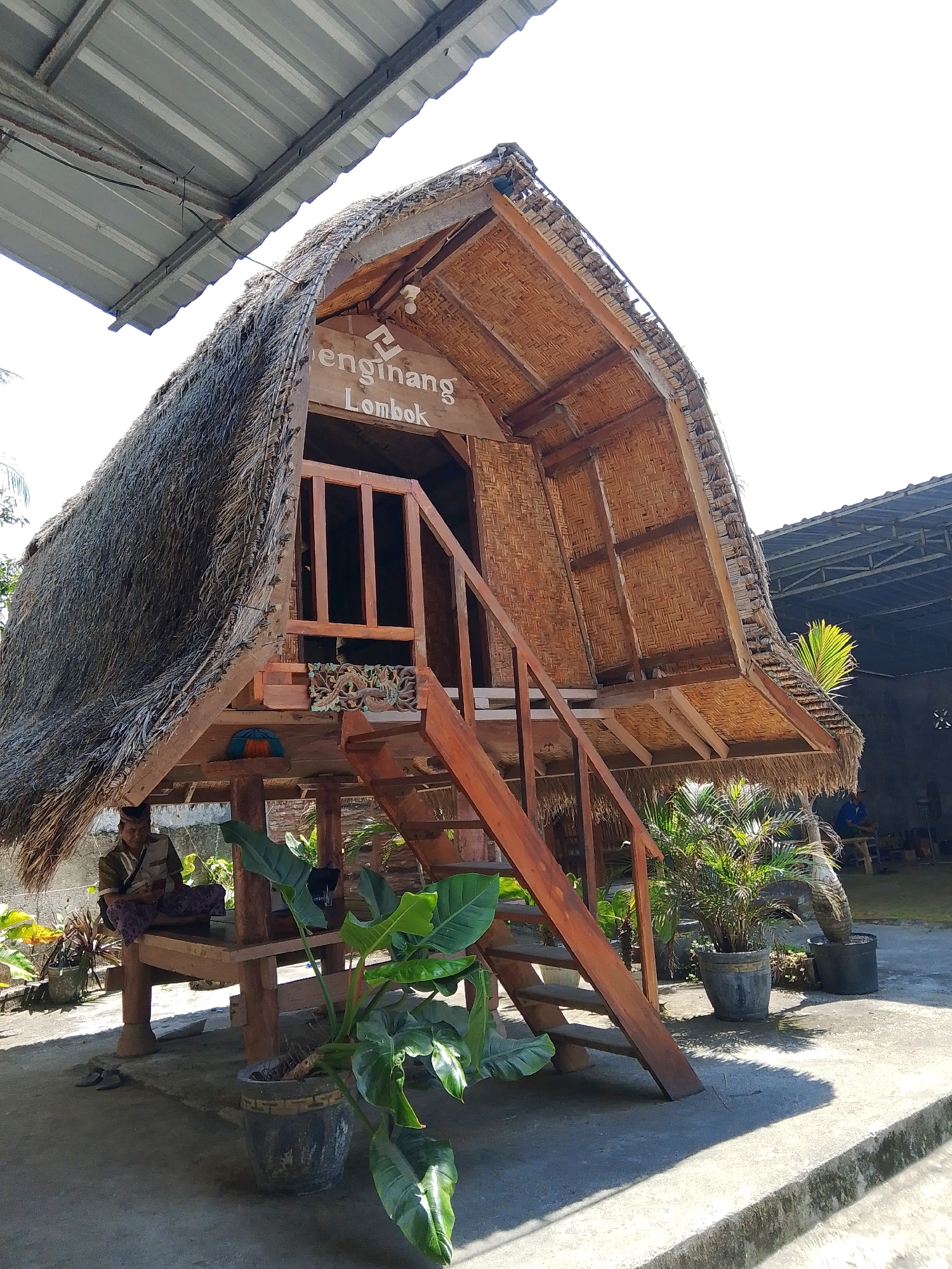 A thatched-roof structure with the words "Enginang Lombok" carved into a wooden sign is the main subject of the image. The structure, typical of traditional Indonesian architecture, is elevated on wooden stilts. A wooden staircase leads up to an open-air upper level covered with woven bamboo. A person, dressed in a traditional sarong and shirt, sits on a wooden bench beneath the structure, looking at a mobile device. Numerous potted plants, including large-leafed varieties and palm trees, are scattered around the concrete ground surrounding the building. In the background, another building with a corrugated metal roof is visible, along with a glimpse of greenery and a person sitting in the distance. The scene appears to be outdoors, under bright sunlight, suggesting a warm climate. The location is identified as Bunkate, Indonesia.
