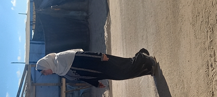 The image, oriented vertically, depicts a person, likely an elderly woman, standing outdoors on a sunny day. She is wearing a white head covering (possibly a hijab or khimar) and a long, dark-colored dress or abaya. The dress features intricate, light-colored embroidered patterns on the sleeves and down the front, suggesting a traditional design. Her hands appear to be held in front of her body, and she is looking downwards or slightly to her right.
The ground beneath her is unpaved, appearing to be dry, light-colored dirt or sand, with visible tire tracks on the right side of the frame. The strong sunlight casts a long, distinct shadow of the woman onto the ground behind her, indicating a bright day, likely late morning or early afternoon.
In the background, there are several makeshift structures. To the left, a wooden frame supports what appears to be a light-colored tarp or fabric roof, with a section of blue tarp visible underneath. Further back and to the right, a darker, possibly black or dark blue, fabric structure, resembling a tent or a tarp-covered shelter, is visible. There is also a rough, light-colored wall or structure, possibly made of concrete or mud brick, in the mid-ground. The sky above is clear and blue with a few wispy white clouds visible in the upper left corner. No discernible text is visible in the image. The overall setting suggests a humble outdoor environment, possibly a rural area or a temporary settlement.