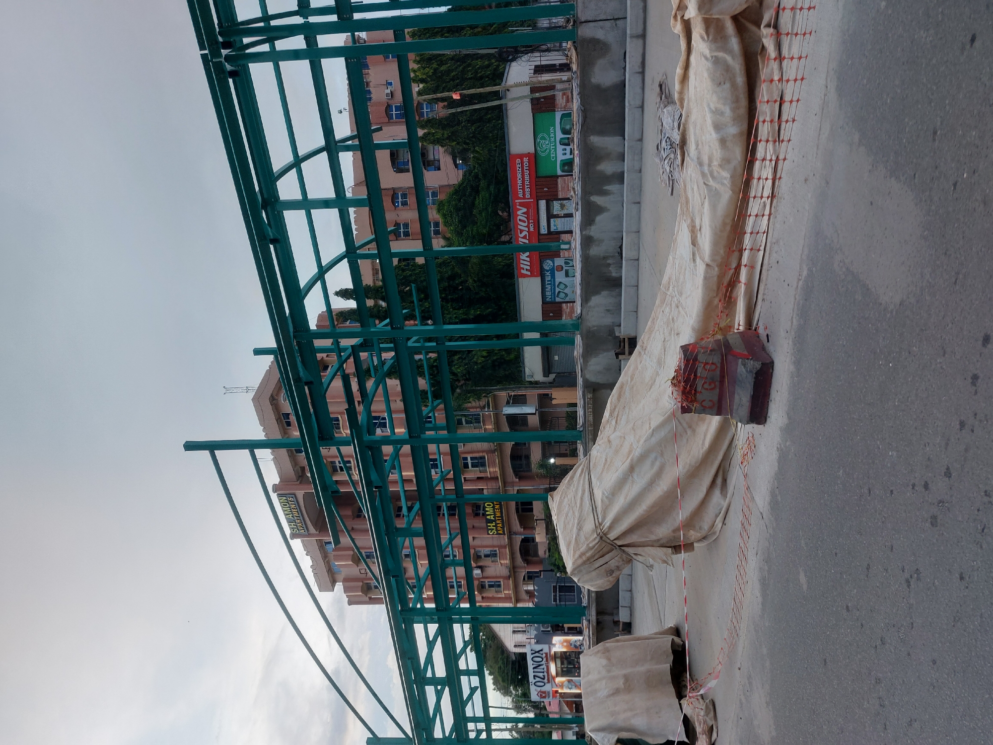 This is an outdoor shot of an urban setting, specifically a street scene in Dar es Salaam, Tanzania. The image has an industrial or construction-site feel.
In the foreground, there is a teal-colored metal structure, potentially a bus shelter or scaffolding under construction. Beside the structure, there are items covered by a light-colored tarp, secured with rope. A red and white barricade tape is also present.
Behind these immediate objects, there is a multi-story building with balconies, identified by a sign that reads "S.H. AMON APARTMENTS". Several business signs are visible, including "HIK VISION AUTHORIZED DISTRIBUTOR" and "ÖZINOX".
The sky is overcast, suggesting it might be early morning or late afternoon. The street is paved, and there is evidence of pedestrian or vehicle traffic.