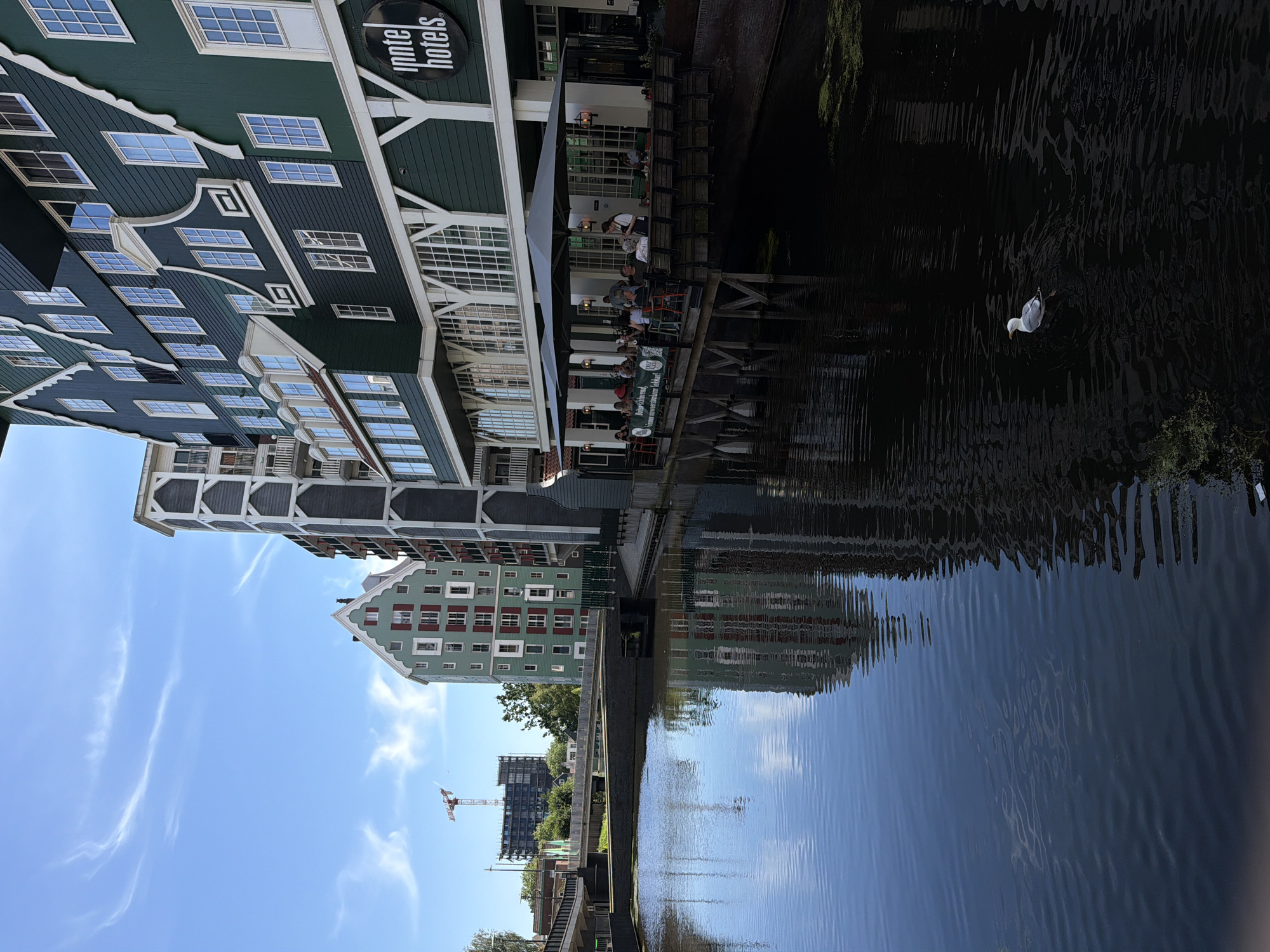 This image depicts the Inntel Hotels, a distinctive waterfront hotel in Zaandam, Netherlands, characterized by its unique facade that mimics traditional Zaanse houses. The hotel building, painted in a deep green, features a stepped gable design and numerous windows. In front of the hotel, an outdoor seating area with large grey umbrellas is set up on a wooden deck extending over the canal. Several patrons are seated at tables, and a server is visible interacting with them. The canal in the foreground reflects the hotel and the blue sky with wispy white clouds. A lone seagull swims in the dark water to the right of the frame. In the background, other multi-story buildings, including a light green one with red shutters, are visible, along with bridges crossing the canal and a construction crane in the distance.
