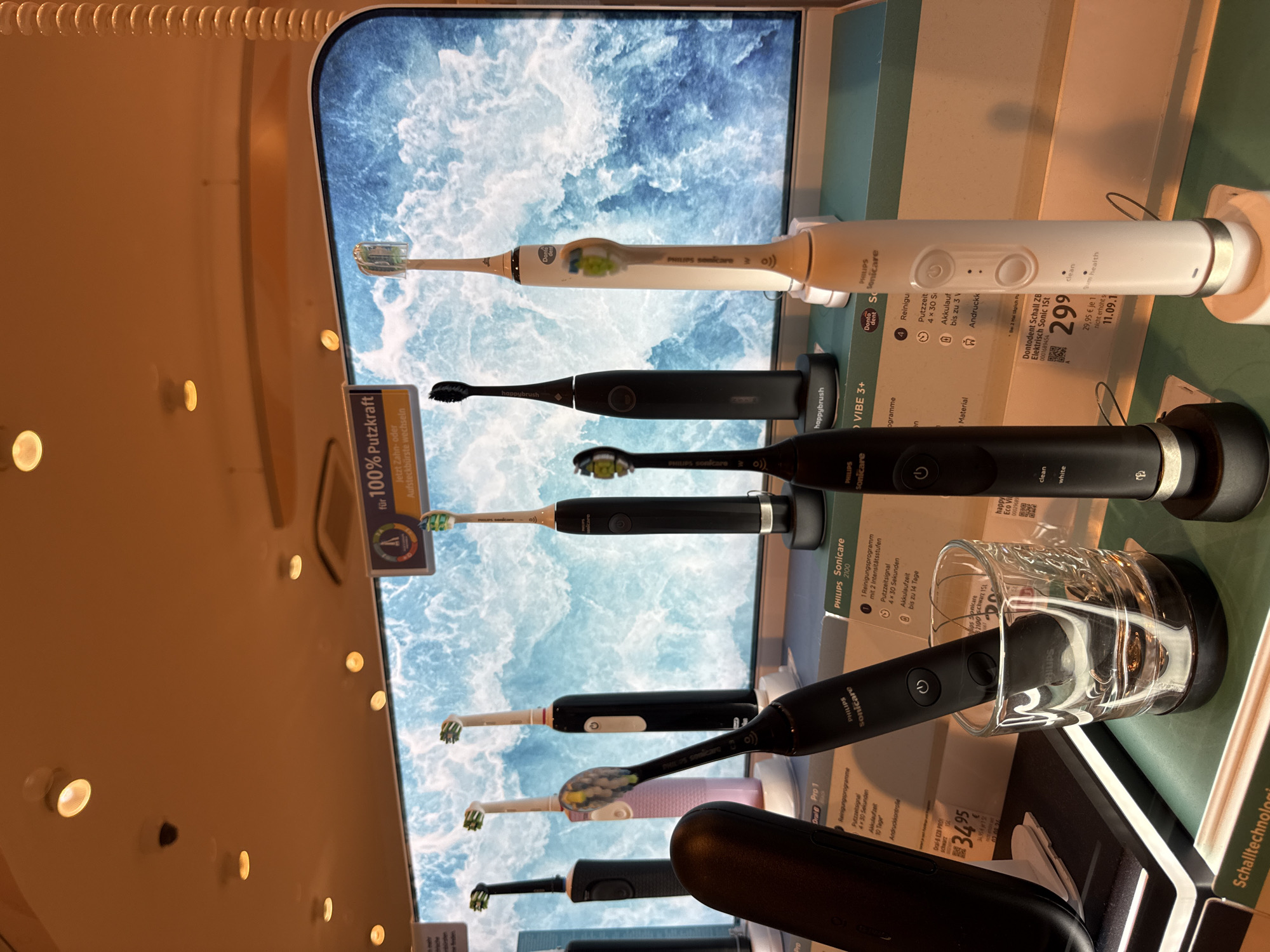 The image shows a retail display of electric toothbrushes. There are multiple Philips Sonicare toothbrushes and at least one happybrush toothbrush visible. The toothbrushes are arranged on shelves against a backdrop that appears to be a digital display showing a seascape. Several price tags and product information labels are also visible. One of the toothbrushes is placed in a clear glass. The lighting suggests an indoor retail environment. The image does not contain any information about the city of Herford, Germany.