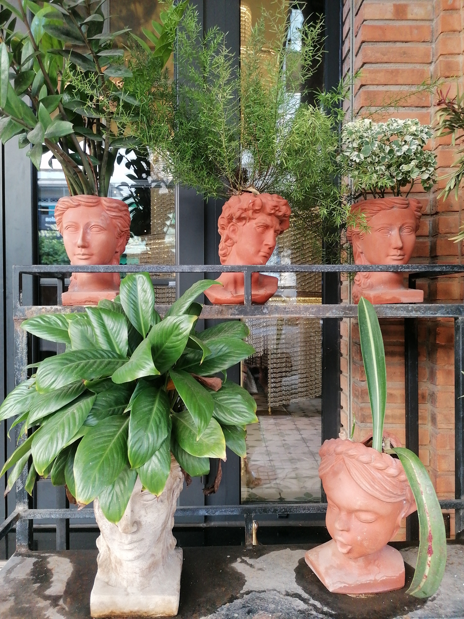 The image shows an outdoor display of plants in head-shaped planters, with the location being Nha Trang Ward, Vietnam. The planters are arranged on a black metal shelf and on the ground. The head-shaped planters on the shelf contain various types of plants with greenery. The planter to the bottom left is a stone white color and holds a large plant with broad green leaves. The planter on the bottom right, on the ground, is a terracotta-colored girl's head, with a couple of leaves sticking out of the top. The background includes a red brick wall and a partially visible building with glass windows. The floor is a dark, mottled surface, appearing slightly wet. There are no people visible in the image.