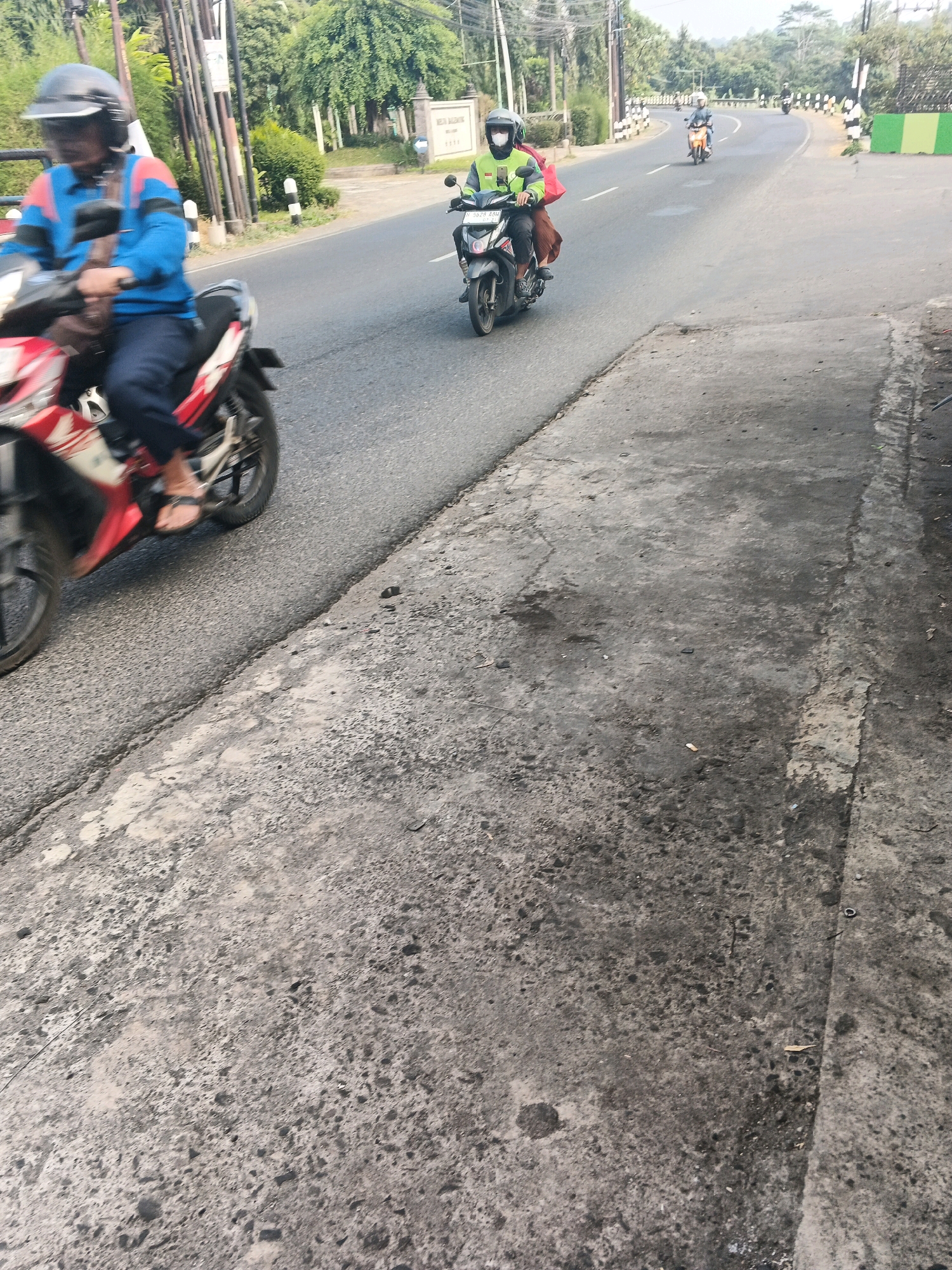 A low-angle, vertical medium shot shows two motorcyclists on a paved road in Ungaran, Indonesia. The motorcyclist in the foreground is wearing a blue and red jacket, a black helmet, and dark pants. They are riding a red and white motorcycle. The motorcyclist in the mid-ground is wearing a green and orange jacket and a white helmet, and is riding a black motorcycle. Further in the distance, another motorcyclist is visible on a light-colored motorcycle. The road has a concrete curb and sidewalk. The background features trees and vegetation. The sky is bright, indicating daytime.