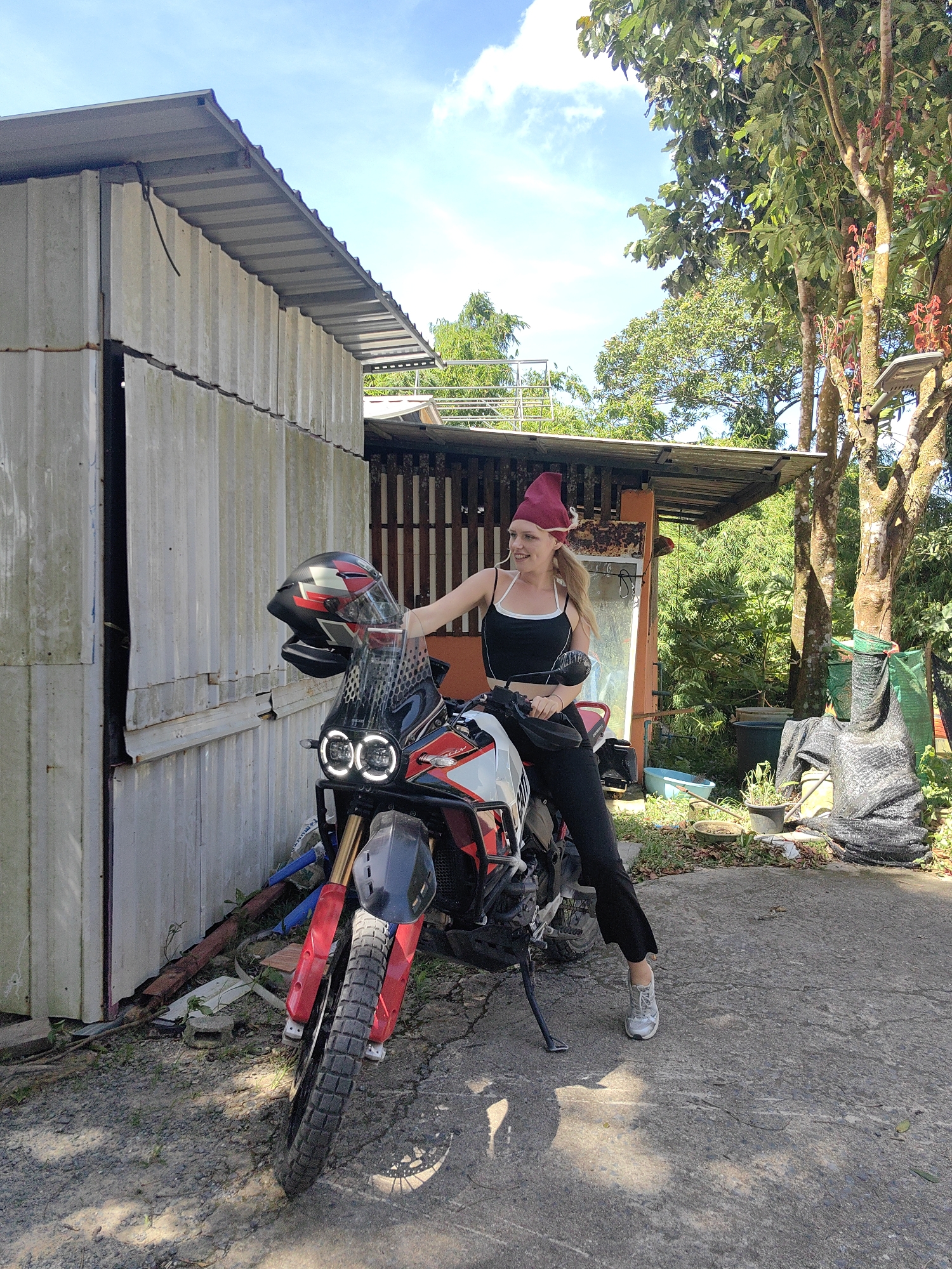 A young blonde woman in a black crop top and black pants, wearing a maroon beanie, sits on a red, white, and black adventure motorcycle. The motorcycle has a large windshield with LED lights. The woman has her right hand resting on the handlebars and is looking to her left with a slight smile. She is standing in a paved outdoor area with gravel and dry leaves on the ground. To her left is a weathered, corrugated metal building. Behind the building, there is a wooden fence and dense green foliage, including a large tree with red flowers. A light blue sky with scattered clouds is visible above. To the right of the motorcycle, there are several potted plants, some dark green mesh bags, and what appears to be part of another building or structure. The scene is lit by bright sunlight, casting shadows on the ground.