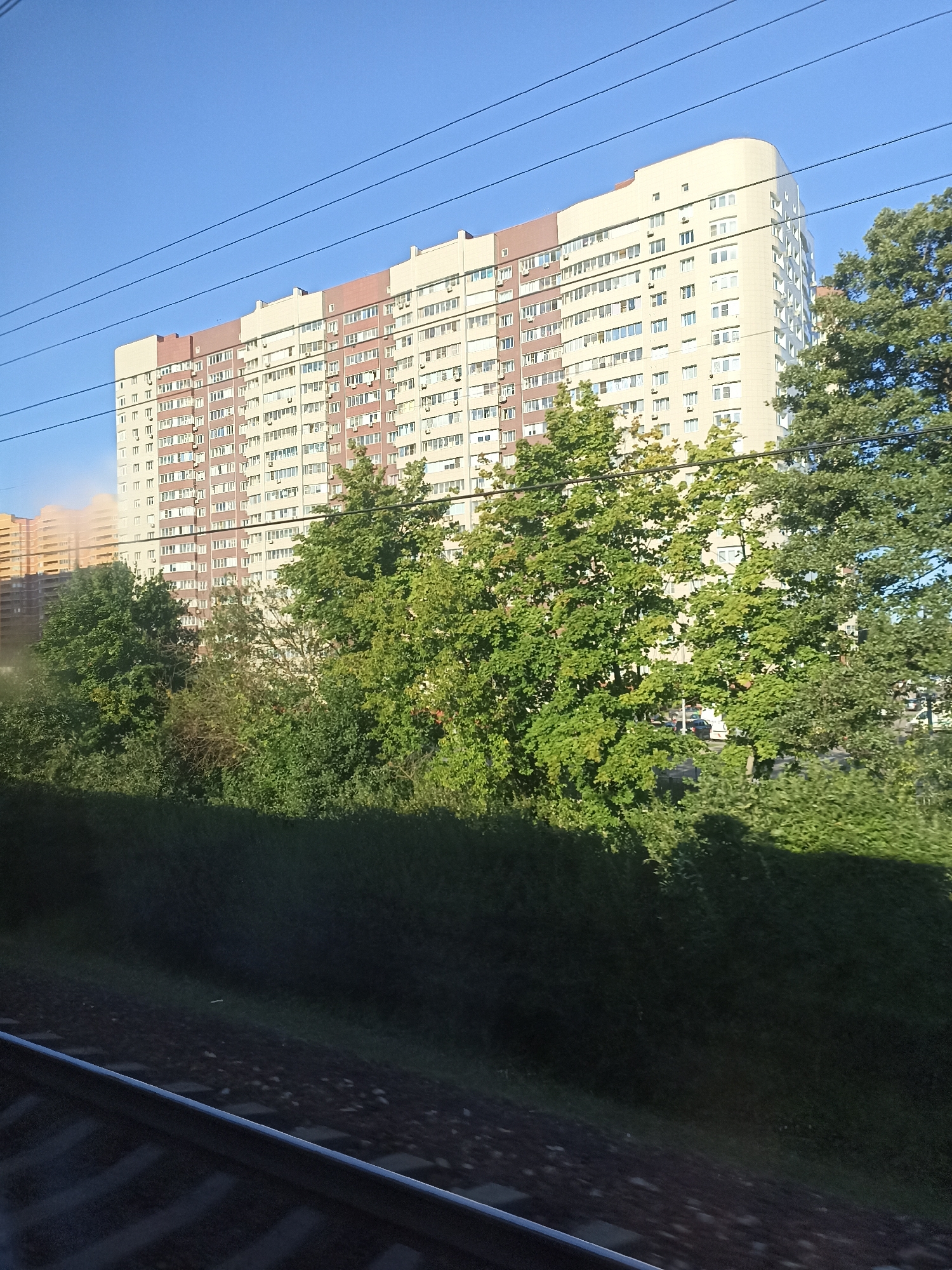 The image shows a large, multi-story apartment building in Shcherbinka, Russia, on a clear day. The building is light beige with reddish-brown accents and numerous windows. In front of the building are several mature green trees. Power lines are visible stretching across the sky. In the foreground, a section of train tracks runs diagonally across the lower portion of the frame, with the dark ballast and rails clearly defined. The lighting suggests it is daytime, with the sun casting shadows on the trees and ground.