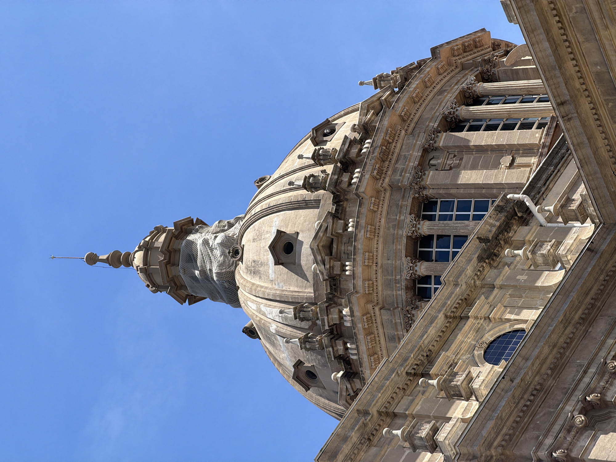 This image shows a close-up, low-angle view of the dome and upper facade of a grand, classical-style building, likely a museum or government building, against a clear blue sky. The architecture features intricate stonework, including cornices, columns with Corinthian capitals, decorative moldings, and a large, segmented dome. The dome itself is topped with a spire. Some sections of the dome appear to be covered with netting, suggesting ongoing maintenance or repair work. The building's facade exhibits a symmetrical design with rows of rectangular windows and smaller, circular or arched windows. The stone is a warm, sandy color, and the sunlight highlights the architectural details. The scene is devoid of people or any overt activity, focusing solely on the imposing structure. This building is the Palau Nacional, located on Montjuïc hill in Barcelona, Spain, overlooking the city.