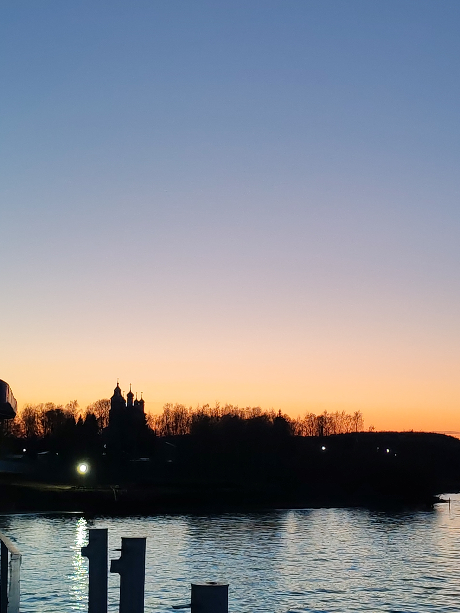 The image captures a serene twilight scene over a body of water, likely a river or lake, with a distant landmass silhouetted against a colorful sky. The sky displays a clear gradient, transitioning from a deep blue at the top, through lighter blues and purples, to warm oranges and yellows near the horizon, indicating either dawn or dusk.
Along the distant horizon, a dense line of trees and possibly elevated land is visible in a dark silhouette. Nestled within these trees, a prominent building with multiple domes and spires, characteristic of a church or monastery, is also silhouetted, adding a distinctive architectural element to the landscape. A single bright, circular light glows on the distant land to the left of the main building, with several smaller, fainter lights visible further along the far bank to the right.
The body of water in the foreground and midground reflects the ambient light, appearing in shades of blue and gray with ripples on its surface. Bright, elongated reflections from unseen light sources or the distant land light shimmer on the water in the immediate foreground, creating vertical streaks of white and light blue. In the bottom foreground, dark, indistinct shapes that appear to be mooring posts or part of a pier structure are visible, partially submerged or rising from the water.
The overall impression is one of tranquility and natural beauty, highlighting the contrast between the vibrant sky and the dark silhouettes of the land and structures. There is no visible text in the image.