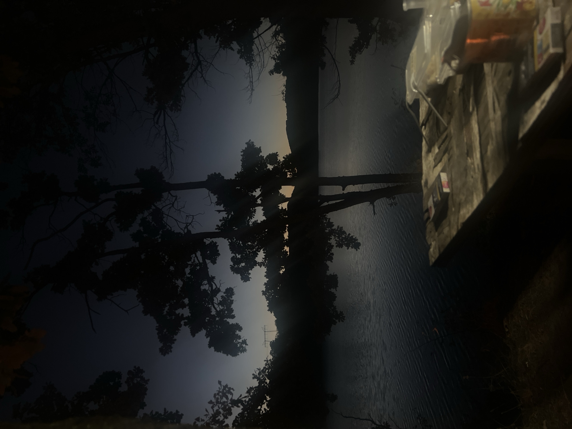 The image depicts a serene lakeside scene at dusk, likely near Brachkovtsi, Bulgaria, given the context. Silhouetted trees frame the tranquil water, reflecting the darkening sky which transitions from a deep blue to a lighter hue near the horizon. In the foreground, a rustic wooden table holds several items, including two packs of cigarettes, a plastic bag containing what appears to be food, and a can. The overall atmosphere is one of quiet solitude, with the natural beauty of the landscape taking center stage. There are no people visible in the image.