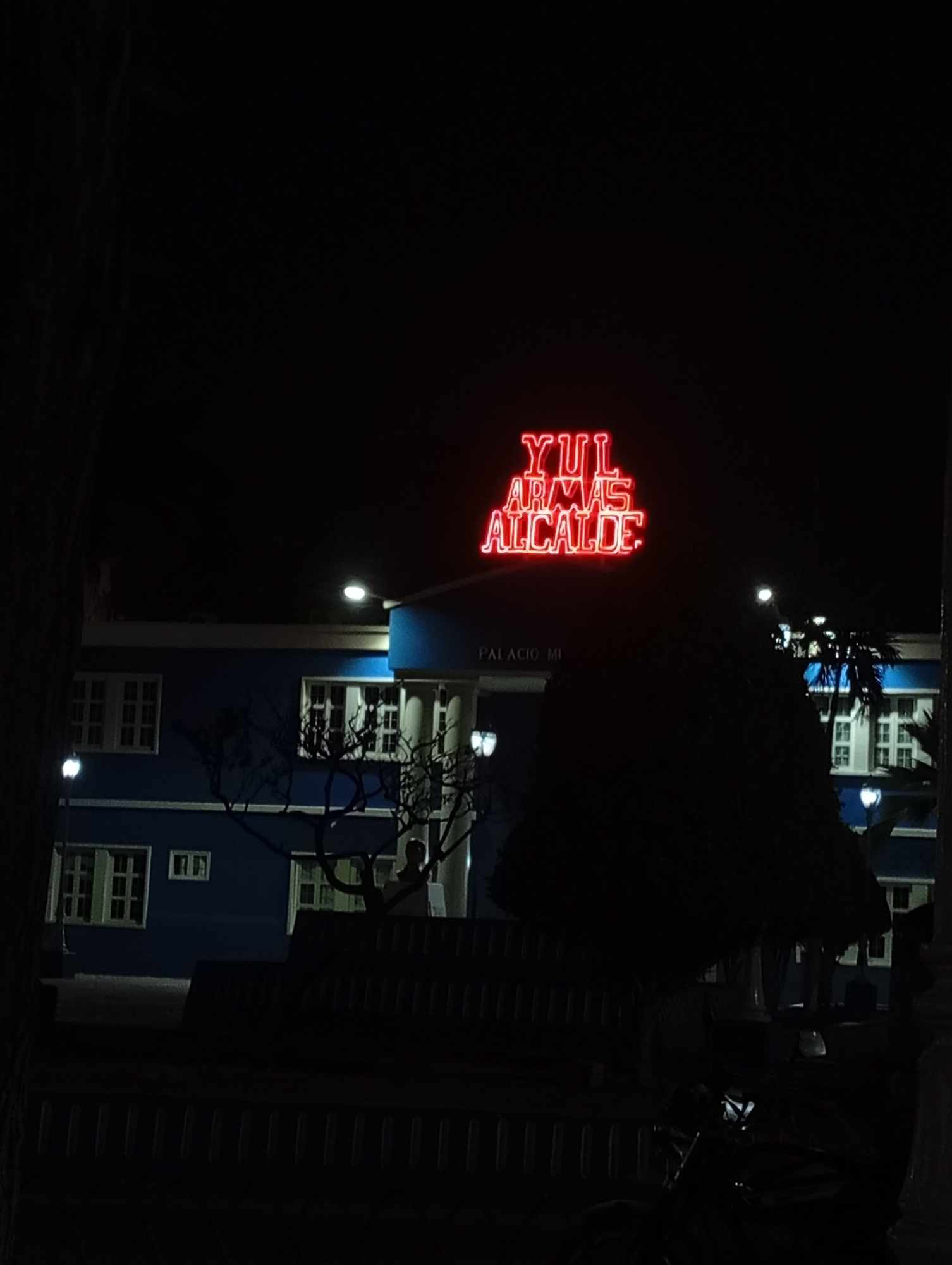 At night, a dark park bench sits in front of the Palacio Municipal in Juan Griego, Venezuela. The building is blue and white, with rows of windows. Above the building, a bright red neon sign reads "YUL ARMAS ALCALDE." Trees are visible in front of the building, obscuring some of its details. Street lamps illuminate the area.