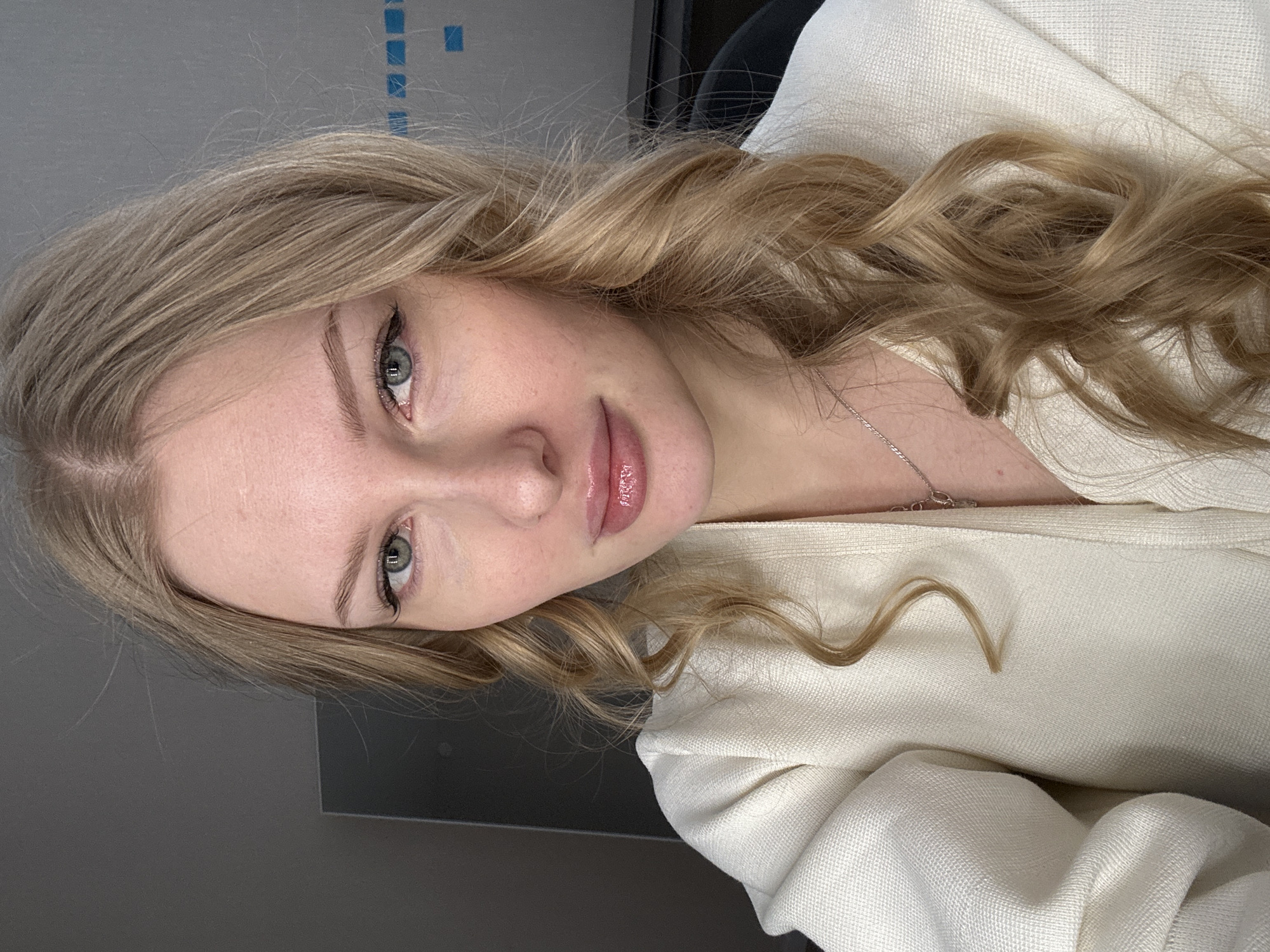A fair-skinned young woman with blonde, wavy hair is positioned at an angle facing the camera. She has light-colored eyes, and her lips appear to have a glossy finish. She is wearing a white or cream-colored textured garment. The background is a neutral gray wall with blue squares visible in the upper left corner. The image does not contain enough information to ascertain the location as Perm, Russia.