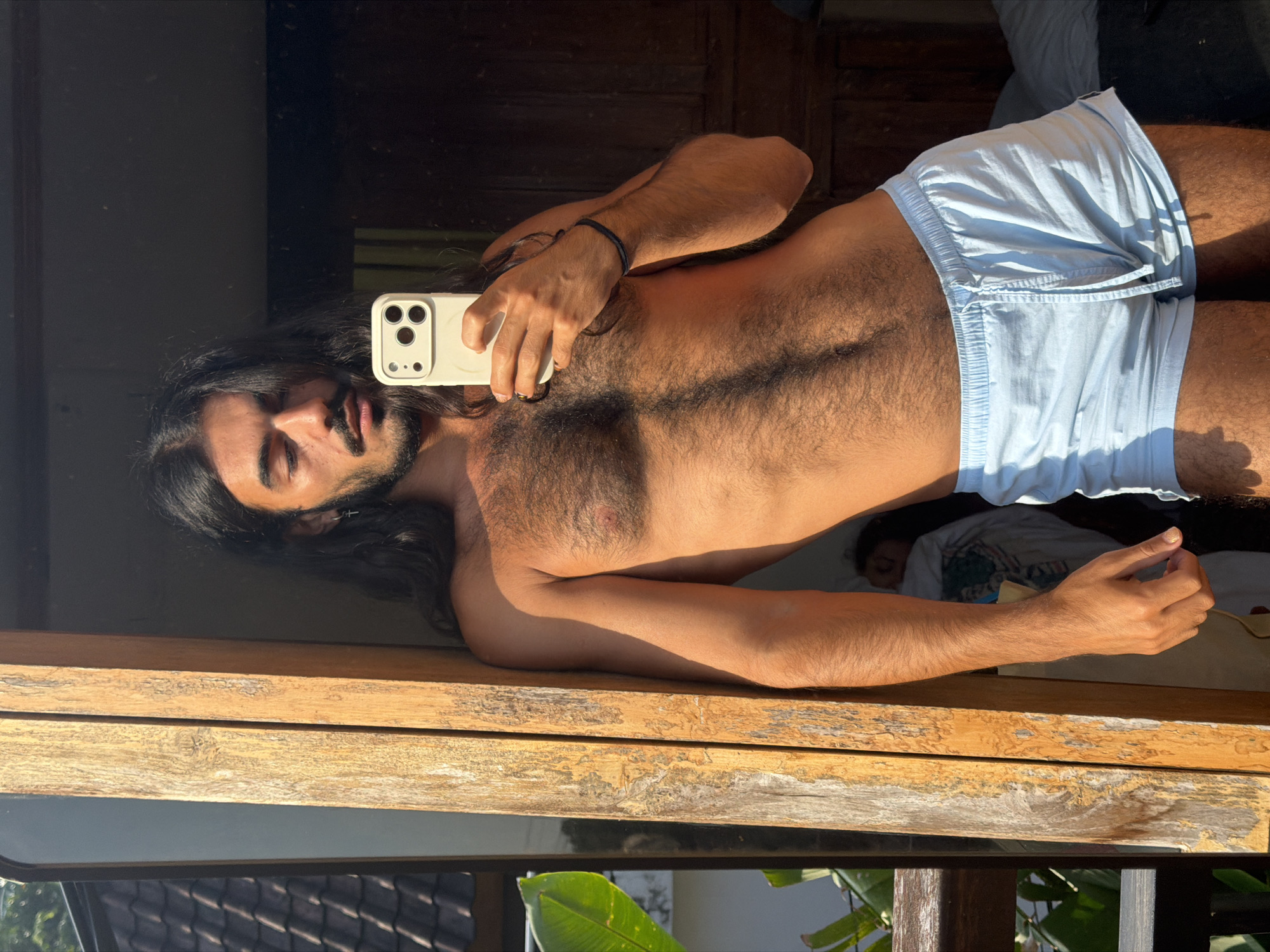 In North Kuta, Indonesia, a person with long dark hair, a mustache, and a beard is reflected in a mirror, taking a selfie with a white smartphone. They are shirtless, exposing a hairy chest and torso, and wearing light blue shorts. The person is leaning against a wooden post, with a window to their left that offers a glimpse of tropical foliage and a tiled roof. The lighting suggests it is daytime, with sunlight casting shadows. In the background, partially visible, another person appears to be sleeping in a bed.