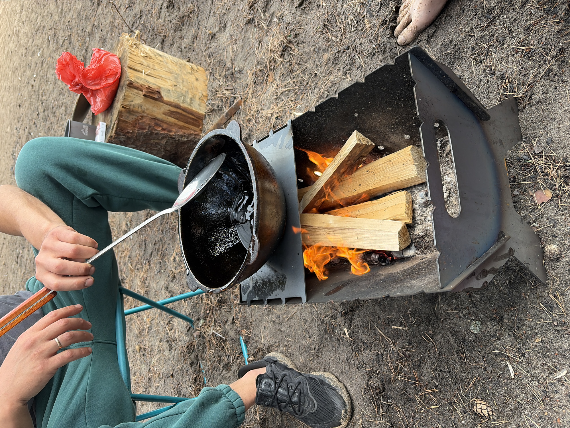 In Vaskelovo, Russia, a person sits on a blue folding chair, wearing green sweatpants and a gray t-shirt. Their hands are visible, with one wearing a ring and holding a metal spatula over a cast-iron pot filled with dark liquid, which is positioned over a fire pit. The fire pit contains burning wood, producing orange flames. To the right of the person, a wooden log serves as a surface for a red plastic bag and a gray cup. The ground is covered with dirt, pine needles, and pinecones. The person is wearing a black sneaker on their left foot. A bare foot is visible on the ground to the right of the fire pit. The overall scene depicts an outdoor cooking activity.