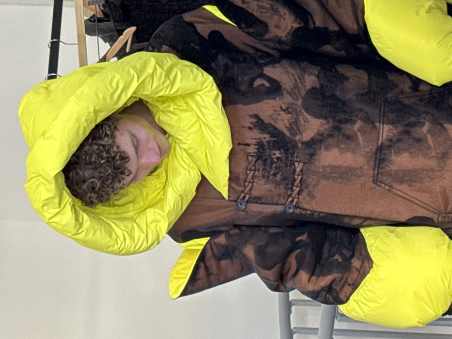 A young male individual with light skin and curly brown hair is depicted in a medium close-up, looking downwards. He is wearing an oversized, hooded garment. The garment features a prominent, voluminous, padded hood in neon yellow, which frames his head. Matching neon yellow padded elements are visible at the cuffs and as angular projections on the shoulders. The main body of the garment is brown with irregular dark brown or black patterns, resembling a bleached or distressed abstract design that includes shadowy figures. Two dark, circular toggle closures with braided fabric loops are present on the chest, above a visible kangaroo-style pocket. In the background, a silver metal railing is partially visible on the left, and a clothing rack holding dark garments on hangers is seen on the right, against a light-colored wall. The scene is located within the city of Milan, Italy, likely in an indoor setting such as a showroom or backstage area.