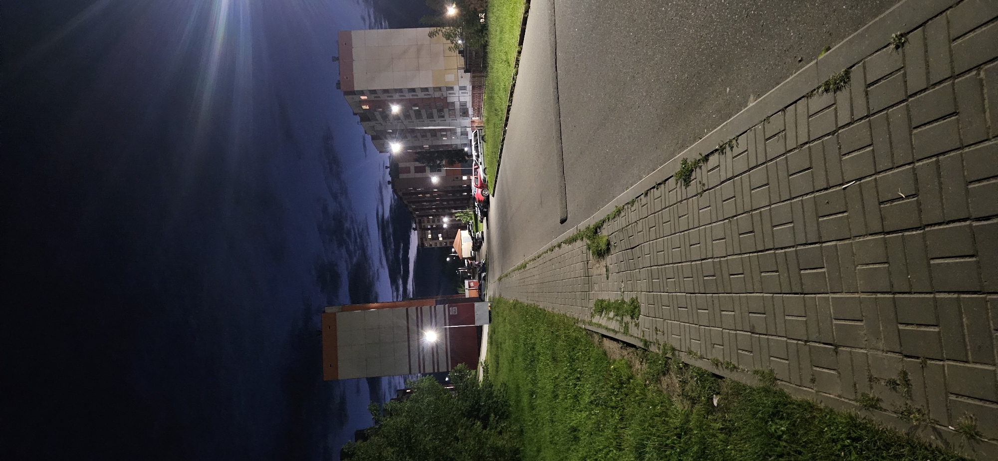The image depicts a night scene in the city of Vitsebsk, Belarus. A paved sidewalk runs along the left side of the frame, bordered by a grassy area and trees. Beyond the grass, a tall, orange and white building is visible. Across a road, a series of multi-story apartment buildings with lit windows stretch into the distance. Several parked cars are scattered in a lot between the road and the apartment buildings. The sky is a dark blue with visible clouds, and streetlights illuminate the scene. The perspective is from ground level, looking down a sidewalk towards the apartment buildings.