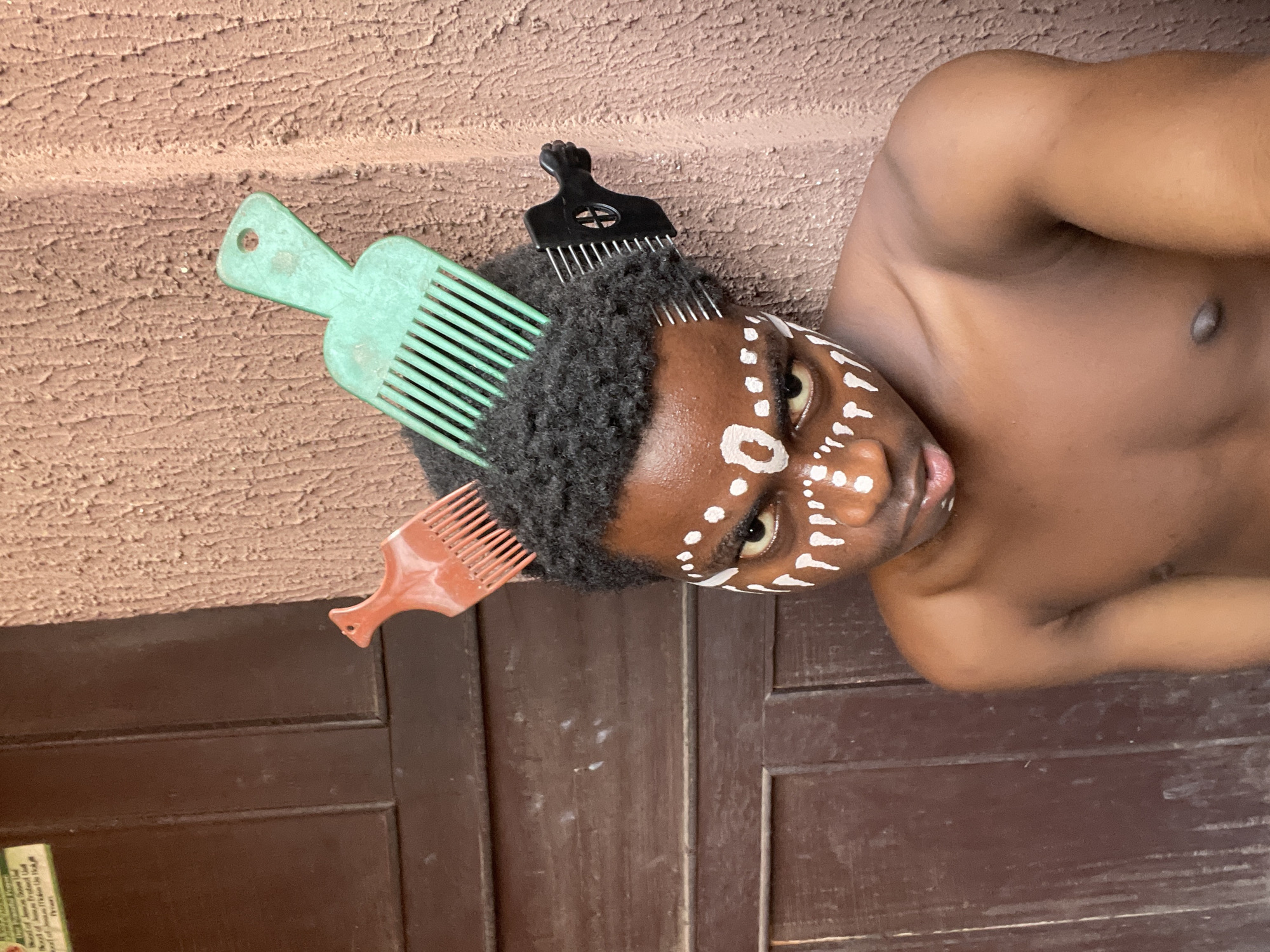 A shirtless person with dark skin and short, curly hair lies on a wooden surface. White paint or makeup marks are applied to their face, resembling tribal-style designs. Three afro picks are inserted into their hair: a green one, a black one, and a brown one. The background shows a textured brown wall and a section of what appears to be a wooden door or panel. The image was captured in Nkpor, Nigeria.
