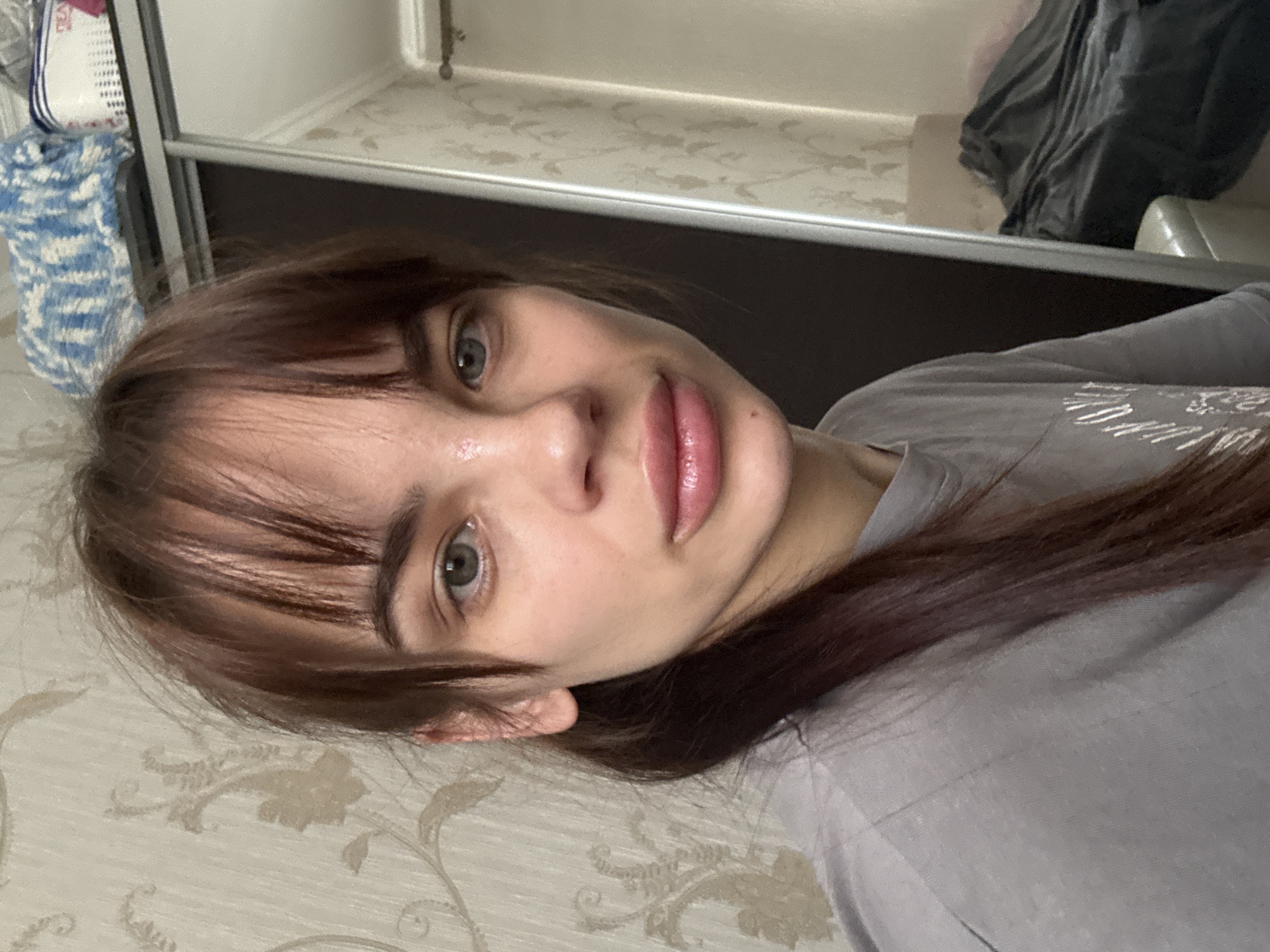 A woman with brown hair and gray-green eyes is seen close-up, lying on what appears to be a patterned surface. She is wearing a gray t-shirt. Behind her, out of focus, there's a wardrobe-like structure with items inside. The location is inside an apartment based on the design and decor. There is no indication of the city Rostov-on-Don.