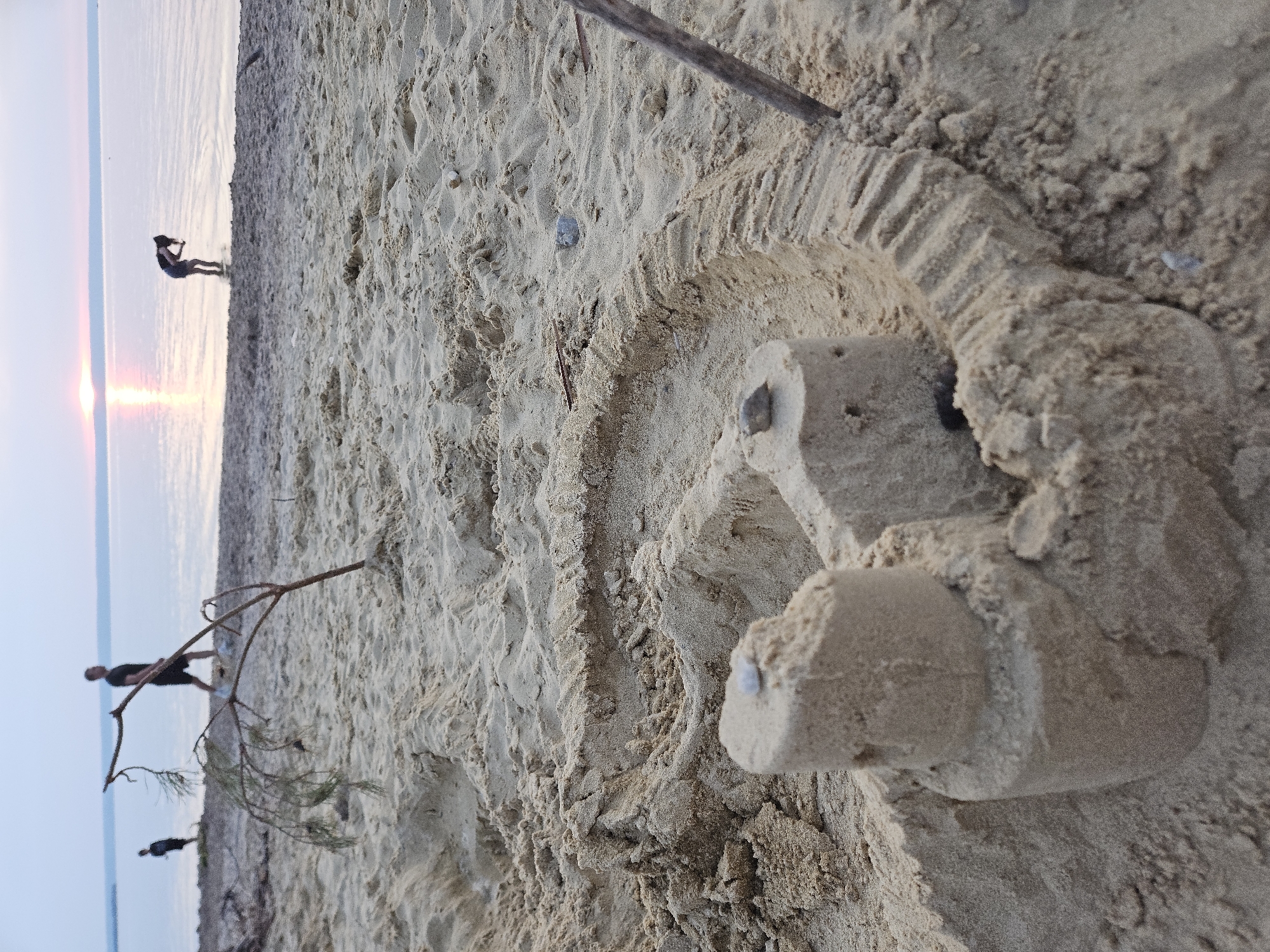 At the sandy shore of Leninsky, Russia, a sandcastle sits in the foreground. The sandcastle is incomplete, with a cylindrical tower and a ramp-like structure extending from it. The texture of the sand is rough and uneven. In the background, the calm, reflective water of a lake or sea stretches to the horizon, where the sky transitions from a light blue to a soft pink and orange, indicating either sunrise or sunset. The sun itself is visible as a bright orb near the horizon, casting a gentle glow on the water. Three individuals are present near the water's edge. One person, a male, is walking away from the viewer. Another individual, also a male, is standing closer to the water. The third person, a female, is bending down, possibly interacting with the water or searching for something on the shore. A thin, dry tree branch with sparse green needles is planted in the sand to the left of the sandcastle. The overall atmosphere is serene and peaceful, capturing a moment of leisure by the water.