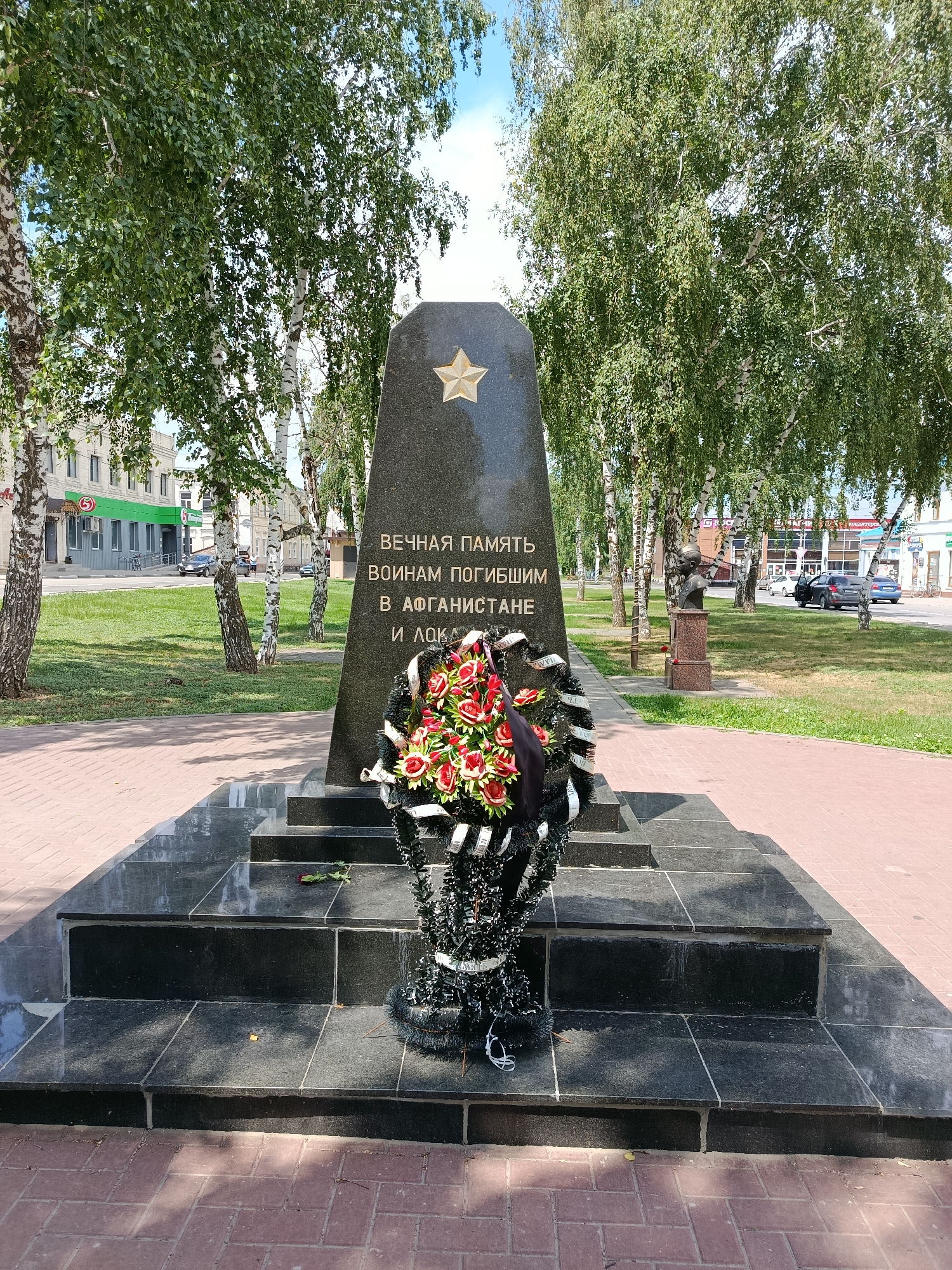 This image depicts a monument in the city of Marx, Russia, dedicated to soldiers who perished in Afghanistan and the local conflict. The monument, made of dark granite, is a tall obelisk topped with a gold star. In front of it, a wreath of red and white flowers with black ribbons is placed on a tiered granite base. The area surrounding the monument is paved with red bricks and a well-maintained lawn. Several birch trees with lush green foliage are visible, along with a bust of a man on a pedestal to the right of the monument. In the background, buildings and a street with passing cars can be seen, indicating an urban setting. The sky is clear and blue, suggesting a daytime scene.