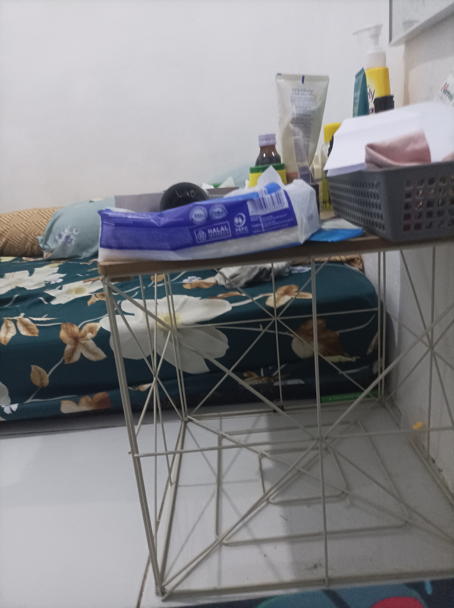 The image depicts an indoor scene with a bed covered in a dark green comforter featuring a floral pattern. A light blue pillow rests on the bed. In the foreground, a white metal shelf unit with a geometric design supports a small wooden shelf. On the wooden shelf, there are various items including a pack of tissues, a dark gray basket containing a pink item, a tube of cream, and a yellow bottle with a pump dispenser. The background shows a white wall and other assorted items, some of which are out of focus. The scene is consistent with a bedroom or living area. It is impossible to determine the specific location within Pamengkang, Indonesia, or any details about people, actions, or interactions as there are none visible in the image.