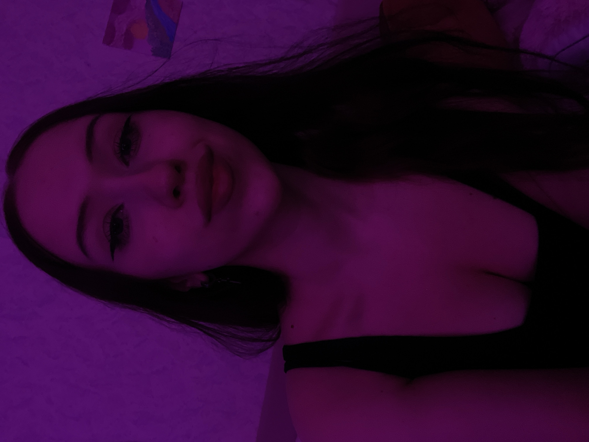 This is a close-up, waist-up, selfie-style photograph of a young woman with long, dark hair. She is wearing a black top and is illuminated by purple lighting. The background appears to be a textured wall with a small abstract painting on the right side. The image is captured in Votkinsk, Russia.