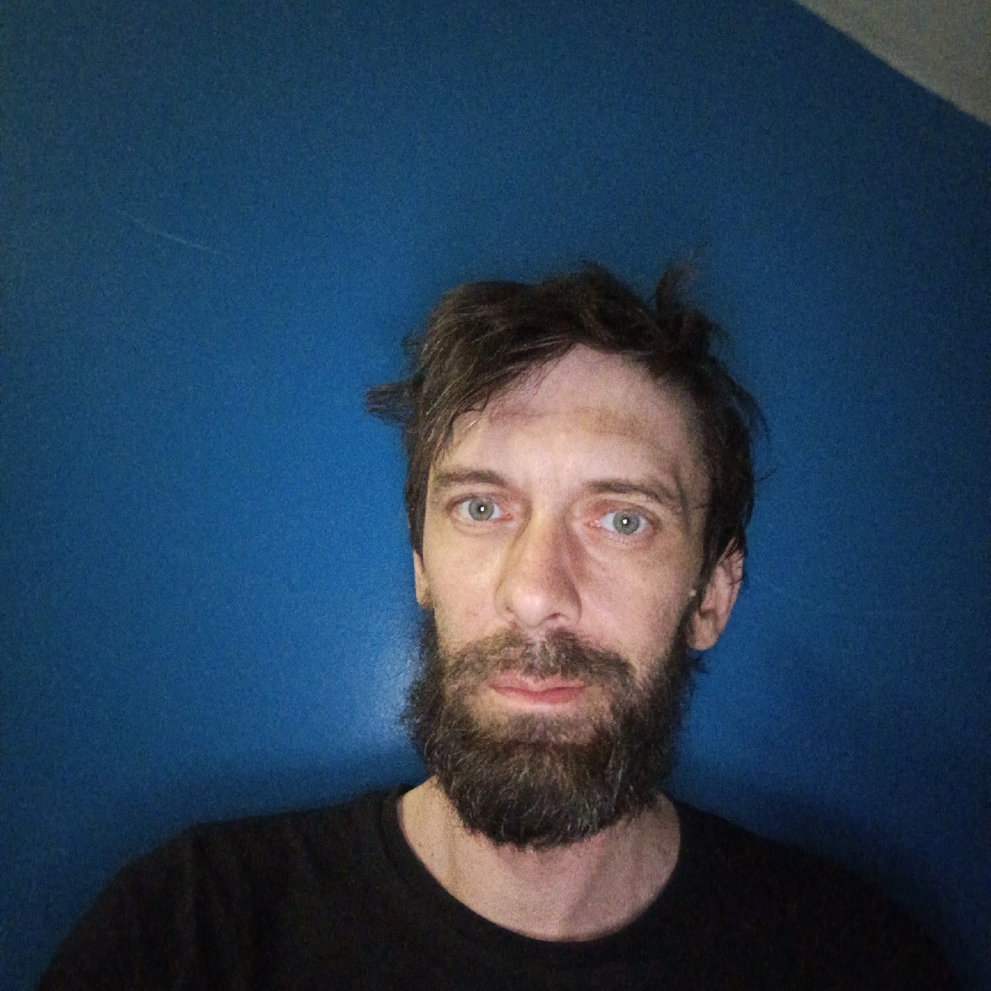 A man with a beard and messy brown hair is facing the camera. He is wearing a black t-shirt and standing in front of a blue wall. The location is Марковский, Russia.