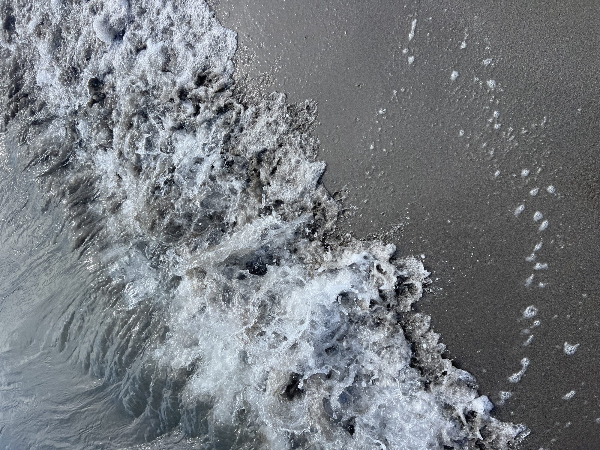 This is a top-down, close-up, outdoor, natural light photograph of a shoreline where the water meets the sand. The image is dominated by the interplay of water and sand, with a wave crashing onto the beach. The water is foamy and turbulent, with white froth and clear droplets. The sand appears dark and wet, with small bubbles scattered across its surface, indicating the receding tide or the movement of water. The composition emphasizes the texture and movement of the water and the granular quality of the sand. There are no visible people, objects, text, or indications of time of day or specific weather conditions beyond what can be inferred from the presence of water and a shoreline. The location is identified as Donji Štoj - Shtoji i Poshtëm, Montenegro.