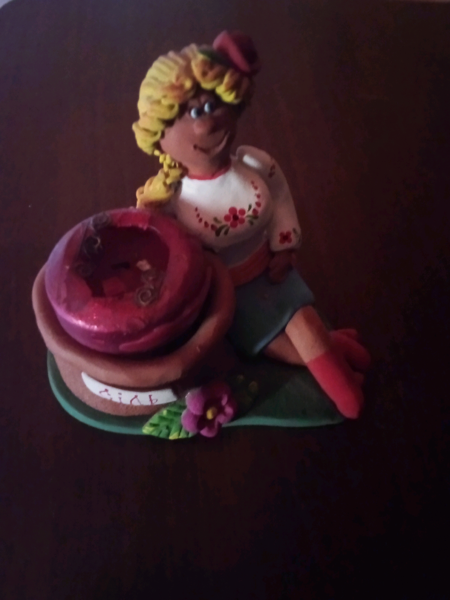 A handmade figurine, likely made of clay or polymer clay, is the sole subject of this dimly lit, eye-level photograph. The figurine depicts a person with tan skin, blonde curly hair tied in a braid, and a red hat. The person is wearing a white top adorned with red floral embroidery and a blue skirt. They are seated with their legs crossed and are leaning on a reddish-brown pot. Inside the pot is a shiny, dark red, bowl-like object with some decorative swirls. A green base with a pink flower and a leaf supports the pot. A white ribbon with what appear to be letters is affixed to the pot. The background is a dark, blurry surface, possibly a wooden table. The lighting is poor, casting shadows and making it difficult to discern finer details. The location in Santomenna, Italy, is not indicated by any visible cues in the image.