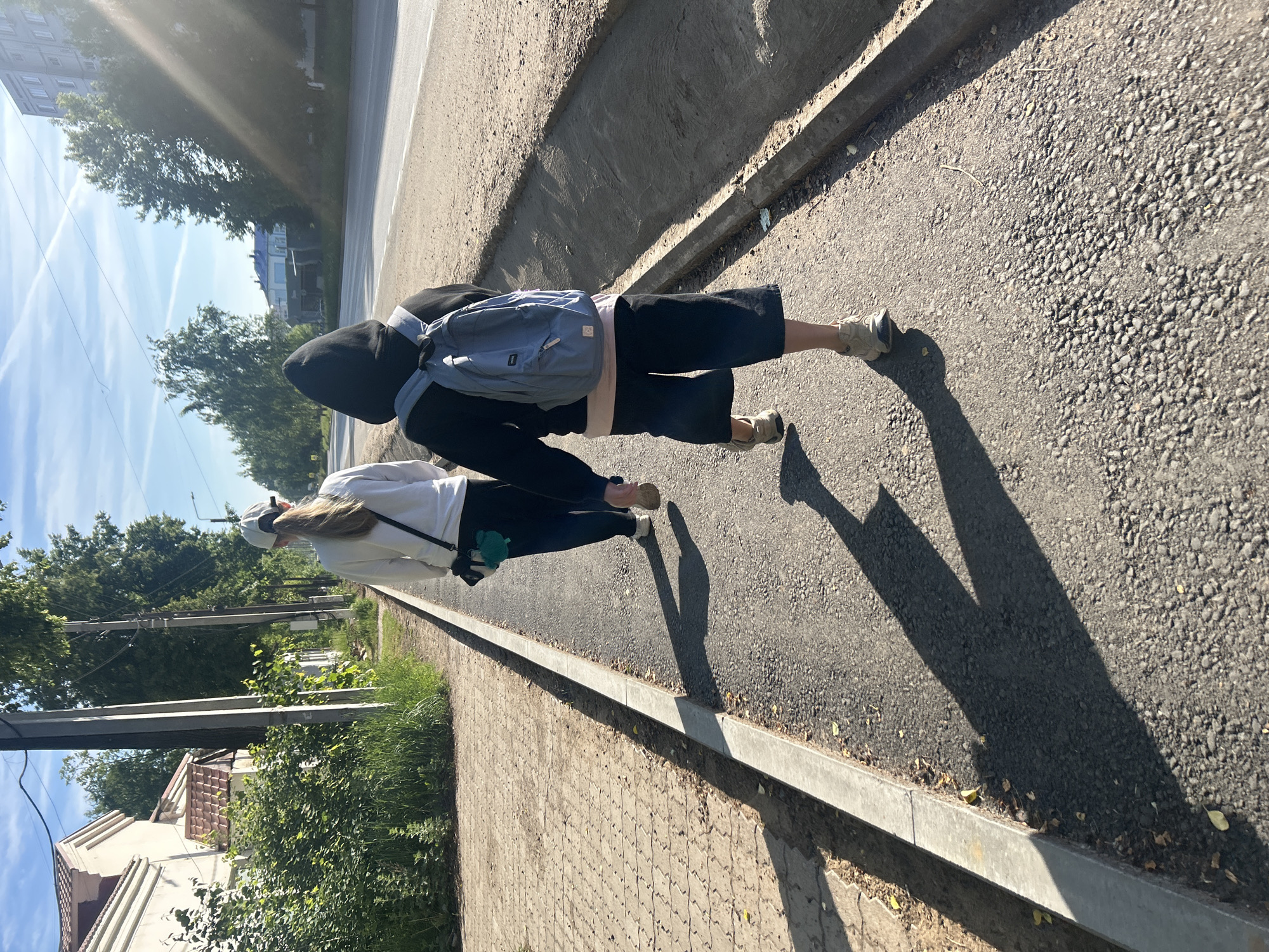 A vertically oriented image captures two individuals walking along a paved surface next to an asphalt road under a bright, sunny sky. The perspective is from slightly above and behind the subjects, looking down the path.
The person in the foreground wears a black hoodie, black shorts, and light-colored sneakers, carrying a light blue backpack. Further along the path, the second person wears a white long-sleeved top, dark pants, and a white baseball cap, with long blonde or light brown hair visible. They are also wearing light-colored shoes and carrying a small, green, fluffy object. Both individuals cast long shadows extending to their left, indicating either early morning or late afternoon sunlight.
To the right of the path is an asphalt road, bordered by a concrete curb. To the left of the walking path is a paved sidewalk made of interlocking bricks. Beyond the sidewalk, there is a strip of grass, bushes, and numerous green trees. In the background, power poles are visible, along with several buildings, including a multi-story apartment block and a single-family house with a brown roof, nestled among the trees. The sky is clear with several faint contrails visible. No text is visible in the image. The scene depicts a sunny day in a suburban or semi-urban environment in Chaikovsky, Russia.