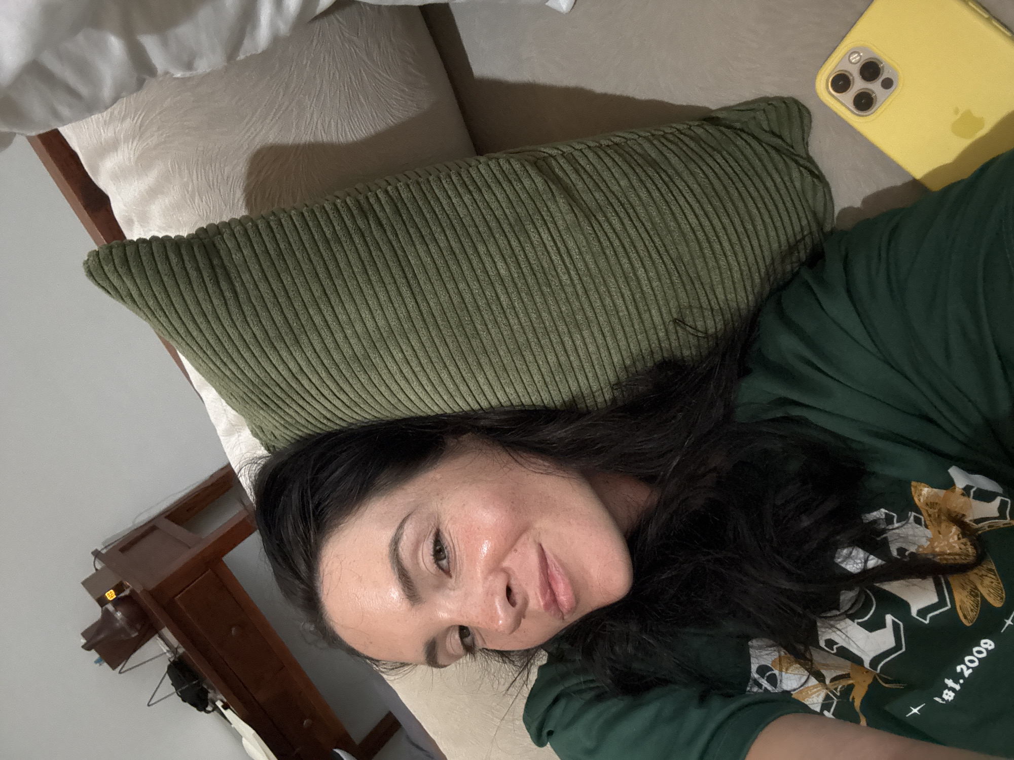 A woman with dark hair lies on a couch, wearing a green t-shirt. She is looking at the camera with a slight smile. To her right, a green corduroy pillow is propped up behind her. Further to the right, a yellow iPhone is visible on the couch. In the background, a wooden desk with drawers and a lamp is situated against a wall. The environment appears to be an indoor residential setting.