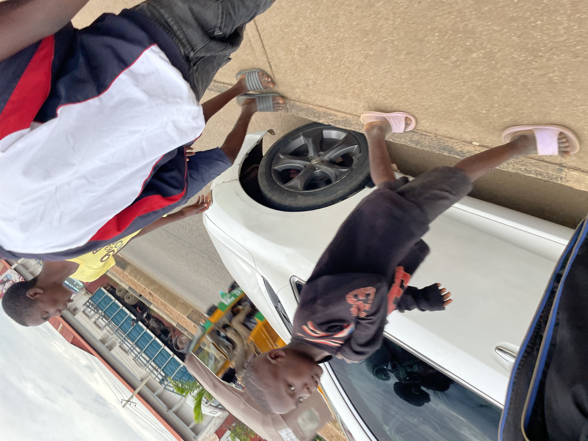 The image depicts an outdoor scene in Barakin Lerawi, Nigeria, characterized by a street with a sidewalk and vehicles. In the foreground, a young boy wearing a black hoodie with red accents and gray shorts walks on the sidewalk, his right leg extended. He is wearing light pink slide sandals. To his left, a white car is parked, and a portion of its front fender is visible. Behind the boy and car, two individuals are walking. One, partially visible on the right, wears a white, navy, and red jacket and dark denim jeans. To the left of this individual, another person in a yellow t-shirt and dark shorts is visible. In the background, a blue truck with a trailer is parked on the street. Adjacent to the truck, a building with a red-tiled roof is visible. A brown tricycle-like vehicle is also present in the middle ground. The environment appears to be overcast, with a cloudy sky.