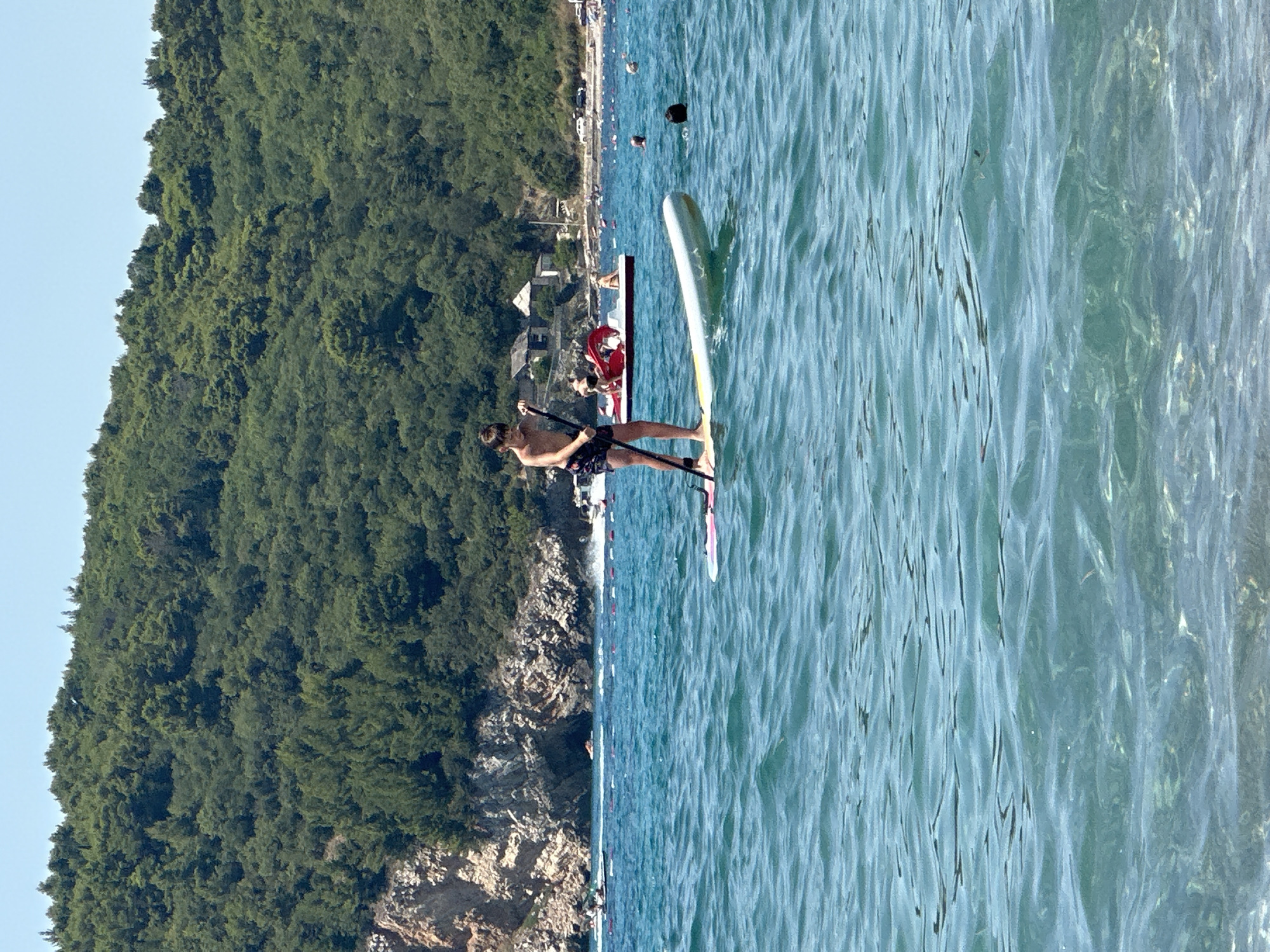 A boy stands on a paddleboard in the turquoise water of the Adriatic Sea, facing left and holding a paddle. He is wearing dark patterned swim shorts. The paddleboard is white with a pink stripe on the front. In the background, a densely forested green hill rises steeply from a rocky coastline. Several buildings are visible nestled amongst the trees on the hillside. Further out in the water, a red pedal boat with people on it is moving away from the shore. Other beachgoers are visible in the distance along a sandy beach. The sky is clear and blue. The scene appears to be a sunny day at a beach in Đurovići, Montenegro.