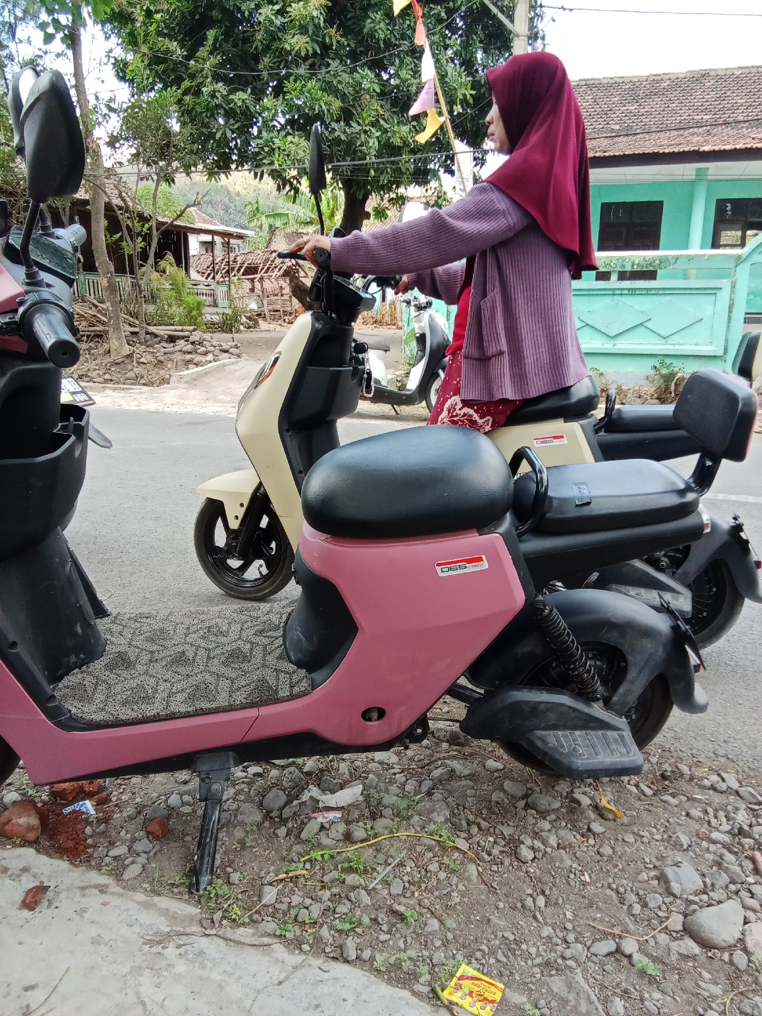 A woman wearing a maroon hijab and a light purple knitted cardigan stands next to a pink electric scooter. She is holding the handlebars of a beige electric scooter parked in front of her. The scooters are parked on a gravelly roadside. In the background, there are trees, a house with a light blue exterior, and a grey tiled roof. The scene appears to be in a rural or suburban area, possibly in the village of Trebungan, Indonesia.