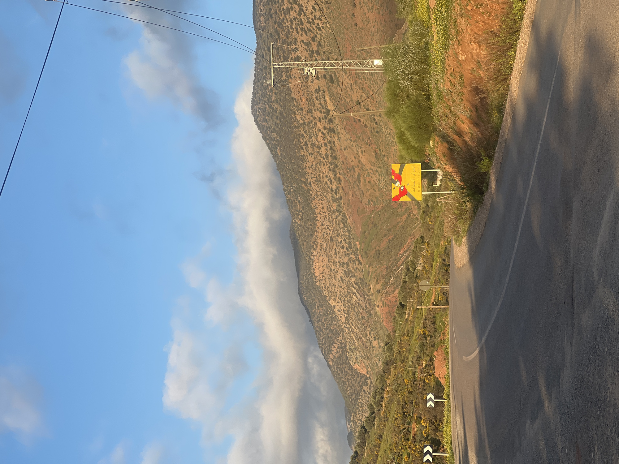 The image shows a road winding along a mountainside in Ouaouizert, Morocco. The road is paved and has a solid white line along the edge. A yellow road sign with red markings is visible next to the road. Utility poles run parallel to the road. The mountainside is covered in sparse vegetation. The sky is blue with scattered clouds.