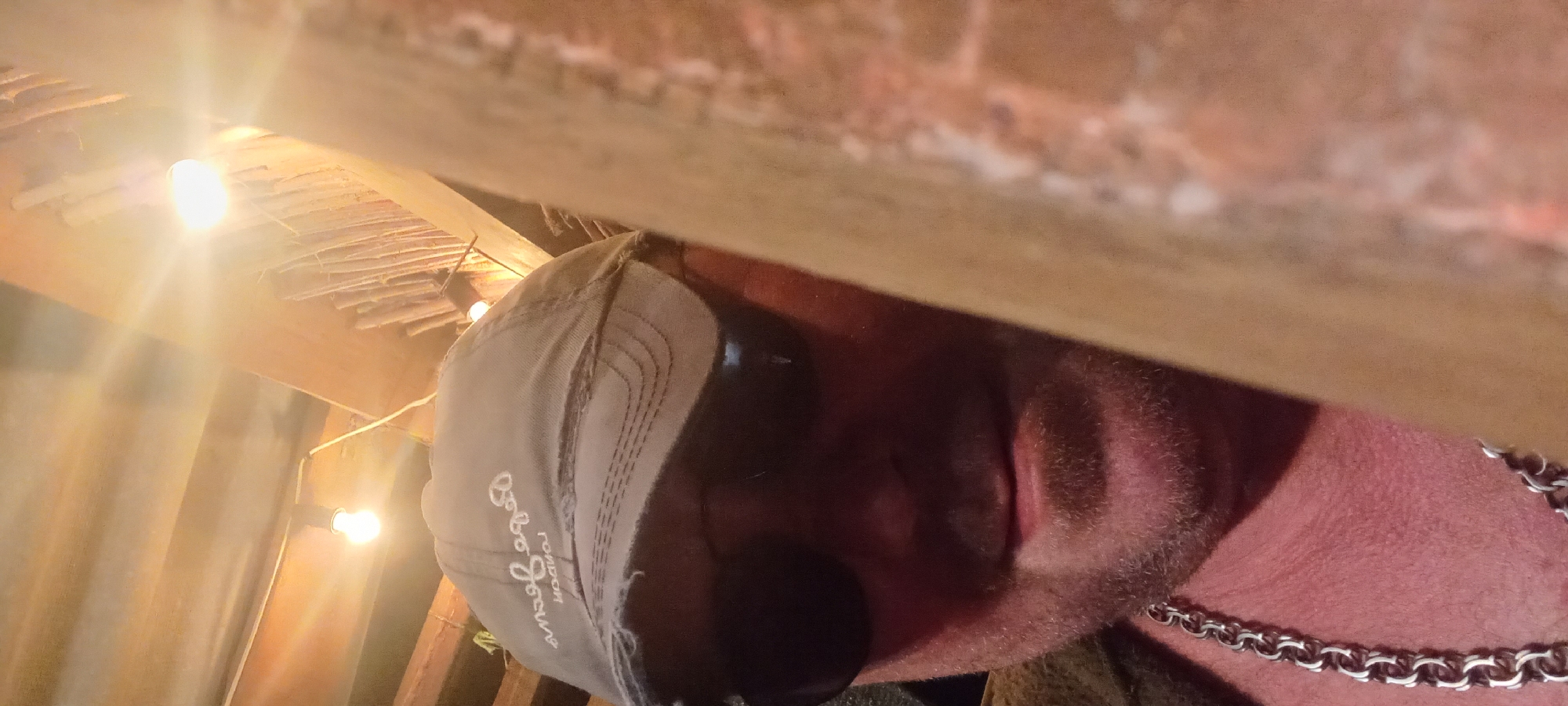 A male subject is depicted in a close-up, indoor selfie. He wears dark sunglasses, obscuring his eyes, and a distressed grey baseball cap with "Pepe Jeans LONDON" inscribed upside down, suggesting a front-facing camera capture. A light stubble or short beard covers his chin and jawline. A heavy silver-colored chain with round links is visible around his neck. The subject's upper body and neck show reddish skin tones, possibly due to lighting or complexion.
The background reveals a rustic interior with exposed wooden beams and a ceiling constructed from corrugated material or planks. Multiple bare incandescent light bulbs, emitting strong, warm light, are visible, creating lens flares in the upper part of the image. One bulb is directly behind the subject's head. A wall or partition to the right of the subject features a vertical arrangement of bundled wooden sticks or reeds, contributing to the rustic ambiance. A large, out-of-focus, light-brown object with vertical textures partially obstructs the right side of the frame, positioned between the subject and the background. The image provides no specific visual indicators to determine the location as Kryvyi Rih, Ukraine.