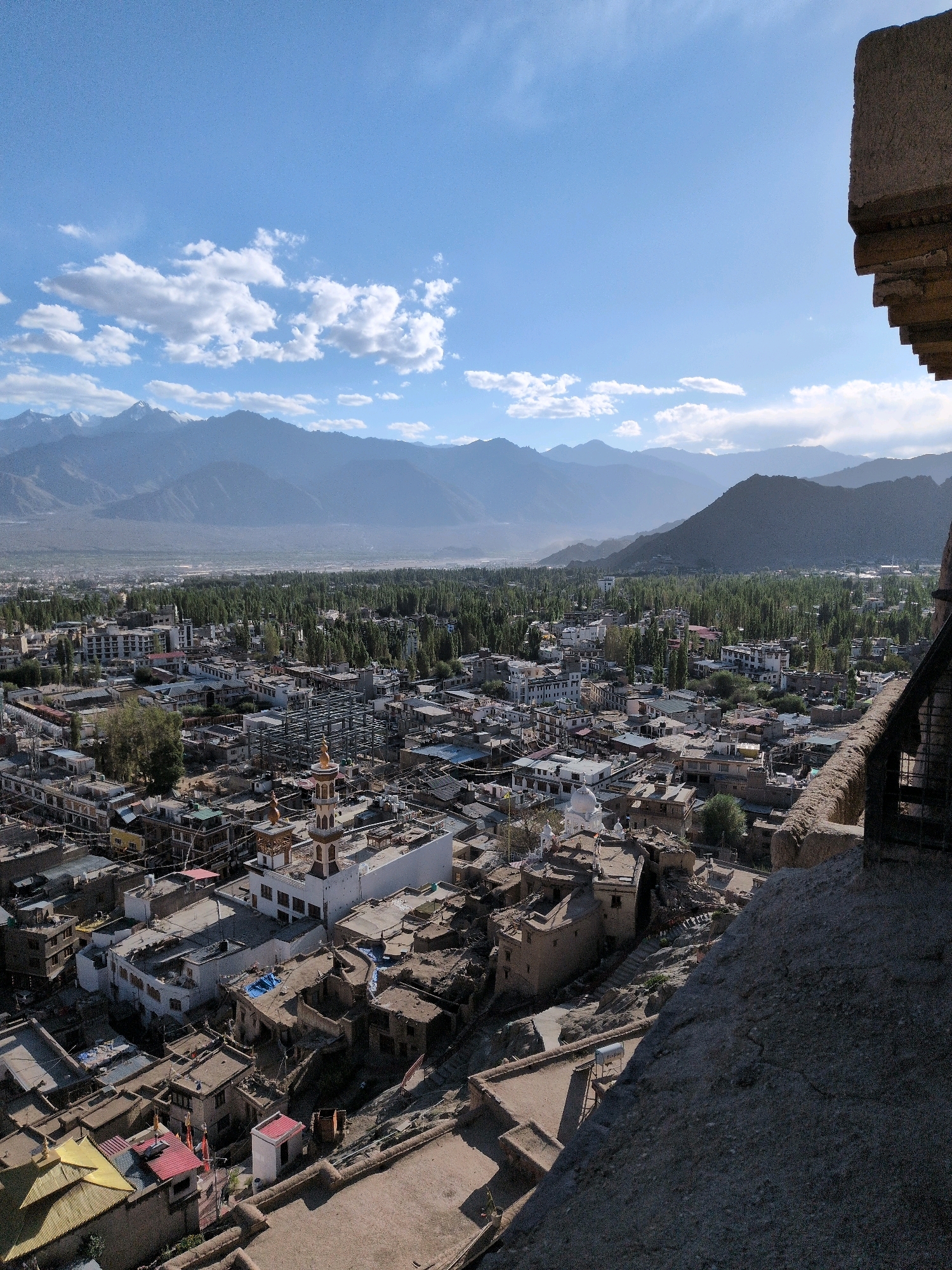 The image is a panoramic view of the city of Leh, India, taken from a high vantage point, likely a palace or monastery. The foreground is dominated by a rough, textured stone wall, part of the structure from which the photo is taken, with a dark, shadowed opening on the right. The middle ground presents a dense urban landscape of buildings with flat roofs, varying in color from white to earthy tones. A prominent white building with a tall, slender tower, likely a mosque, stands out in the mid-section of the city. Interspersed among the buildings are patches of lush green trees, forming a dense canopy that stretches towards the background. The background is characterized by a wide expanse of mountains with jagged peaks, fading into a hazy, atmospheric distance under a bright blue sky dotted with white clouds. The sunlight casts shadows and illuminates different parts of the city and the natural landscape, creating a sense of depth and scale.
