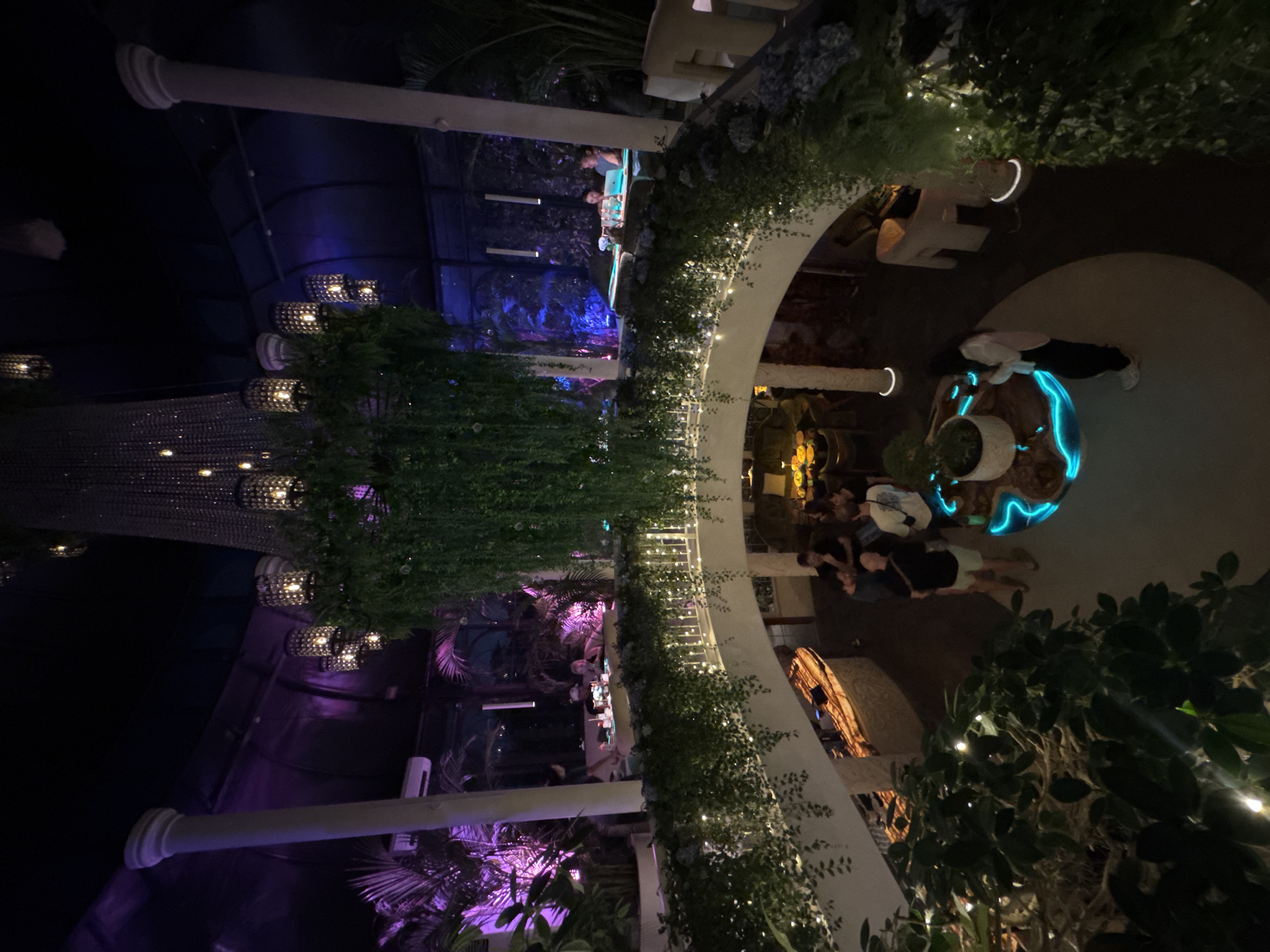 The image depicts the interior of a multi-level restaurant or bar in North Kuta, Indonesia. The space is elaborately decorated with lush greenery, including hanging plants and potted trees, creating a tropical ambiance. Large, white pillars support the structure, and a grand, ornate chandelier hangs from the ceiling, adorned with smaller, lantern-like light fixtures. The lighting is atmospheric, with purple and blue hues cast throughout the space, highlighting the plants and architectural features.
On the upper level, several people are seated at tables, some appearing to be dining or socializing. On the lower level, a group of individuals are gathered, with some standing and others seated around a central, illuminated table with a circular, glowing blue design. A woman in dark pants and a light-colored jacket is standing near this table, looking at her phone. A large, light-colored counter with an illuminated wooden surface is also visible on this level. The overall scene suggests a lively and stylish establishment designed for evening entertainment.