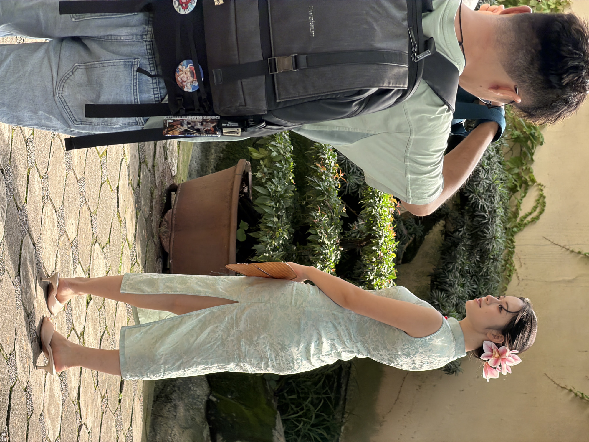 In Yuzhong District, China, a young woman in a light blue, sleeveless qipao with floral patterns and a high neckline, adorned with a pink flower in her hair, poses for a photograph. She holds a light brown clutch in her left hand. She wears light-colored mule heels and her left leg is visible through the slit in her qipao. To her left, a person wearing a light green t-shirt and a large black backpack is taking her photo with a camera. The backpack is adorned with several pins and tags. The person is wearing light blue denim jeans. They are standing on a paved walkway made of irregular stone tiles. Behind them, lush green bushes and plants are visible, with a large brown pot to the left of the woman. A textured wall forms the background. The lighting suggests it is daytime, with shadows indicating sunlight.