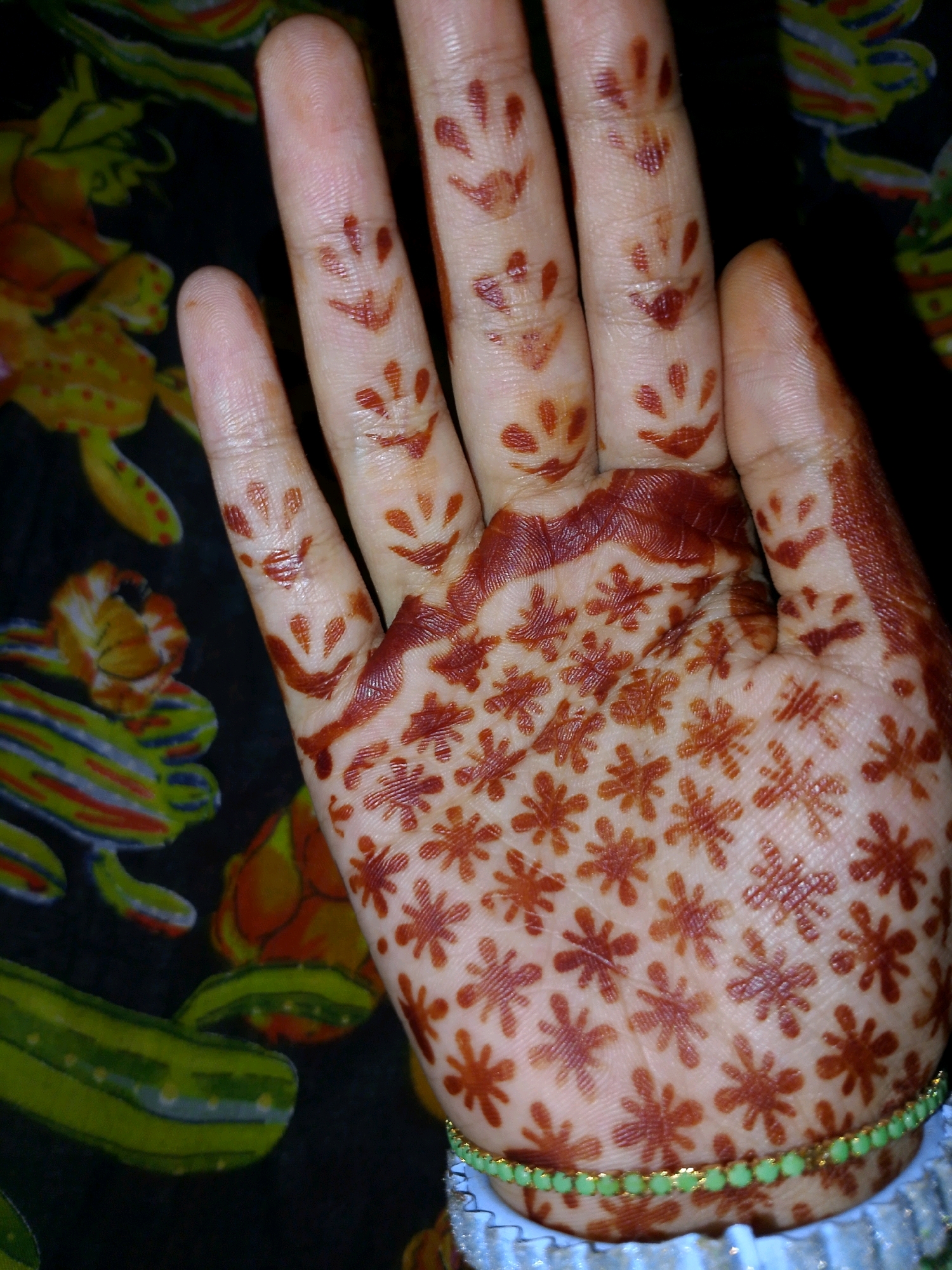 A close-up shot captures a hand adorned with henna designs. The henna, a reddish-brown dye, is intricately applied in floral patterns across the palm and fingers. Some fingers also feature simple smiley face designs. The background is a dark fabric with a colorful, abstract pattern that appears to be a textile. A beaded green bracelet is visible on the wrist. The image does not provide any information about the city of Thane or any specific location within it.