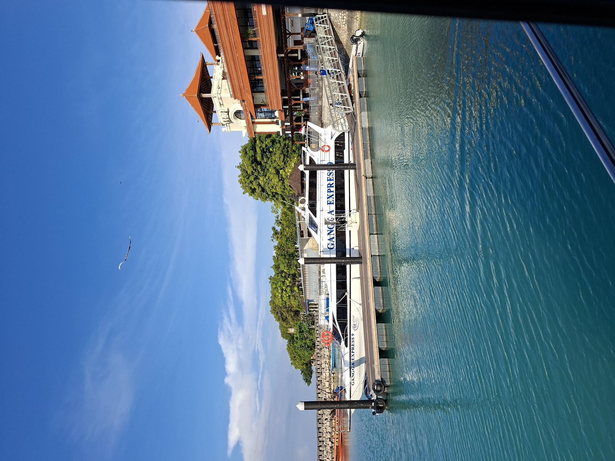 The image shows a clear blue sky with some wispy clouds and a kite flying. Below, a bright turquoise body of water is calm, with slight ripples. A white passenger boat, named "GANGGA EXPRESS 9," is docked at a wooden pier. The boat has several windows and life preservers visible. To the right of the boat and pier, a multi-story building with a distinctive orange tiled roof and balconies stands, suggesting a tropical resort or hotel. Lush green trees are visible behind the building and along the coastline. In the distance, a rocky breakwater extends into the water. The scene appears to be a harbor or marina in Nusa Penida, Indonesia, likely on a sunny day.