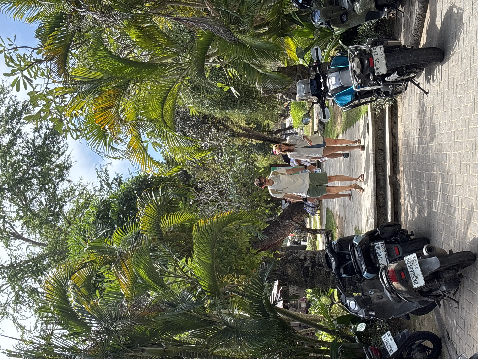 In Kuta Selatan, Indonesia, a paved walkway lined with lush tropical foliage leads into a shaded area. Two individuals, a man wearing a white t-shirt and olive green shorts and a woman in a light-colored open shirt and denim shorts, are walking away from the camera down the path. Several motorcycles are parked on the right side of the walkway, including a bright blue dual-sport motorcycle and a dark gray scooter. On the left side, more motorcycles are visible, along with some traditional stone carvings and umbrellas suggesting a commercial or tourist area. The environment is characterized by dense green palm trees and other tropical plants, with a bright sky visible through the canopy.