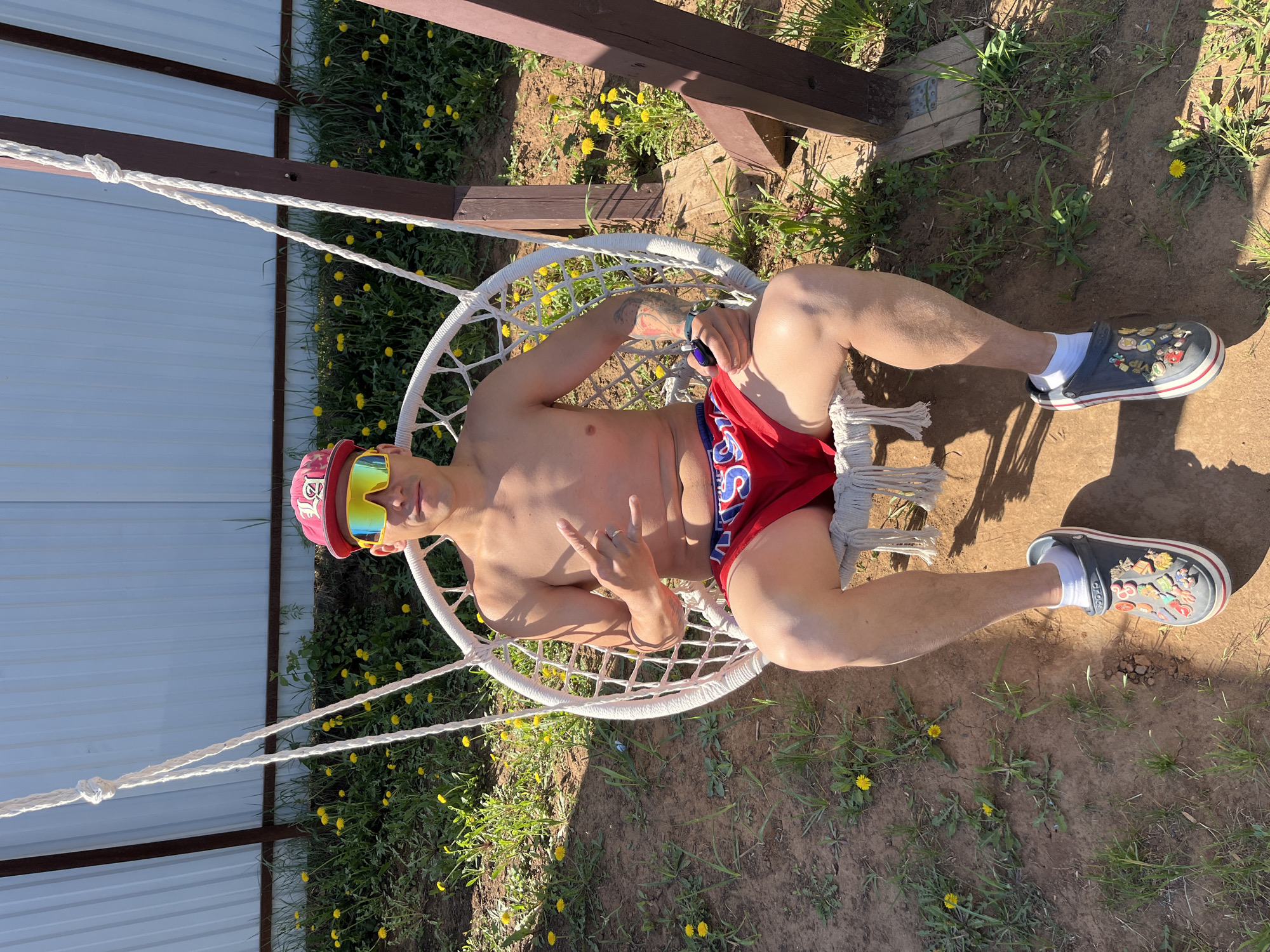 A shirtless man wearing red swim trunks, a red cap, and yellow-tinted sunglasses is lying in a white macrame hanging chair. He is wearing Crocs shoes with charms. The location is outdoors in a grassy area with dandelions. A fence and wooden structure are in the background. The city of Chaikovsky, Russia is not discernible from the image.